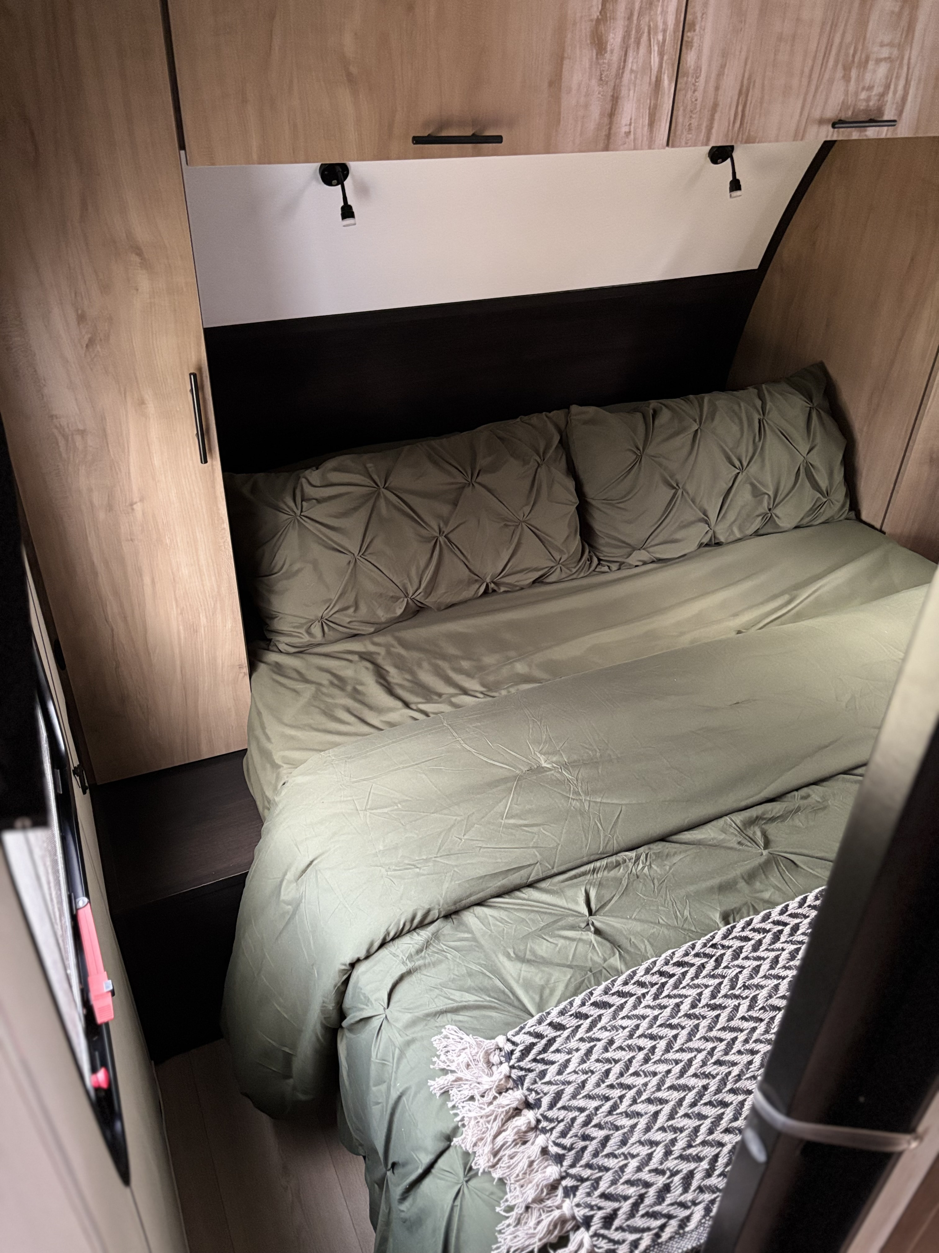 Interior of an RV rental: a cozy bedroom with olive green bedding, wood-look cabinets, and reading lamps.. Forest River Cherokee Black Label 2025