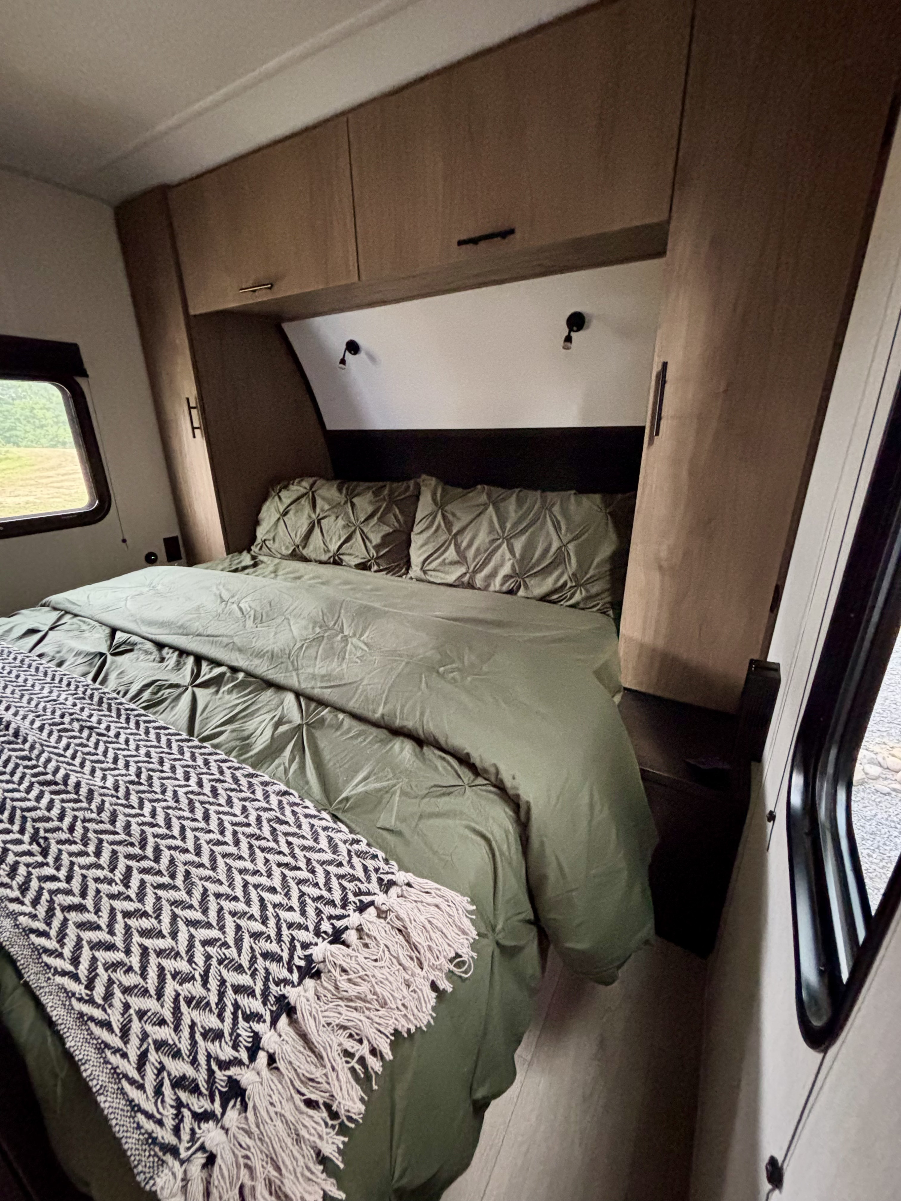 Cozy RV rental bedroom featuring an olive green bed with a patterned throw, overhead storage cabinets, and bright windows.. Forest River Cherokee Black Label 2025