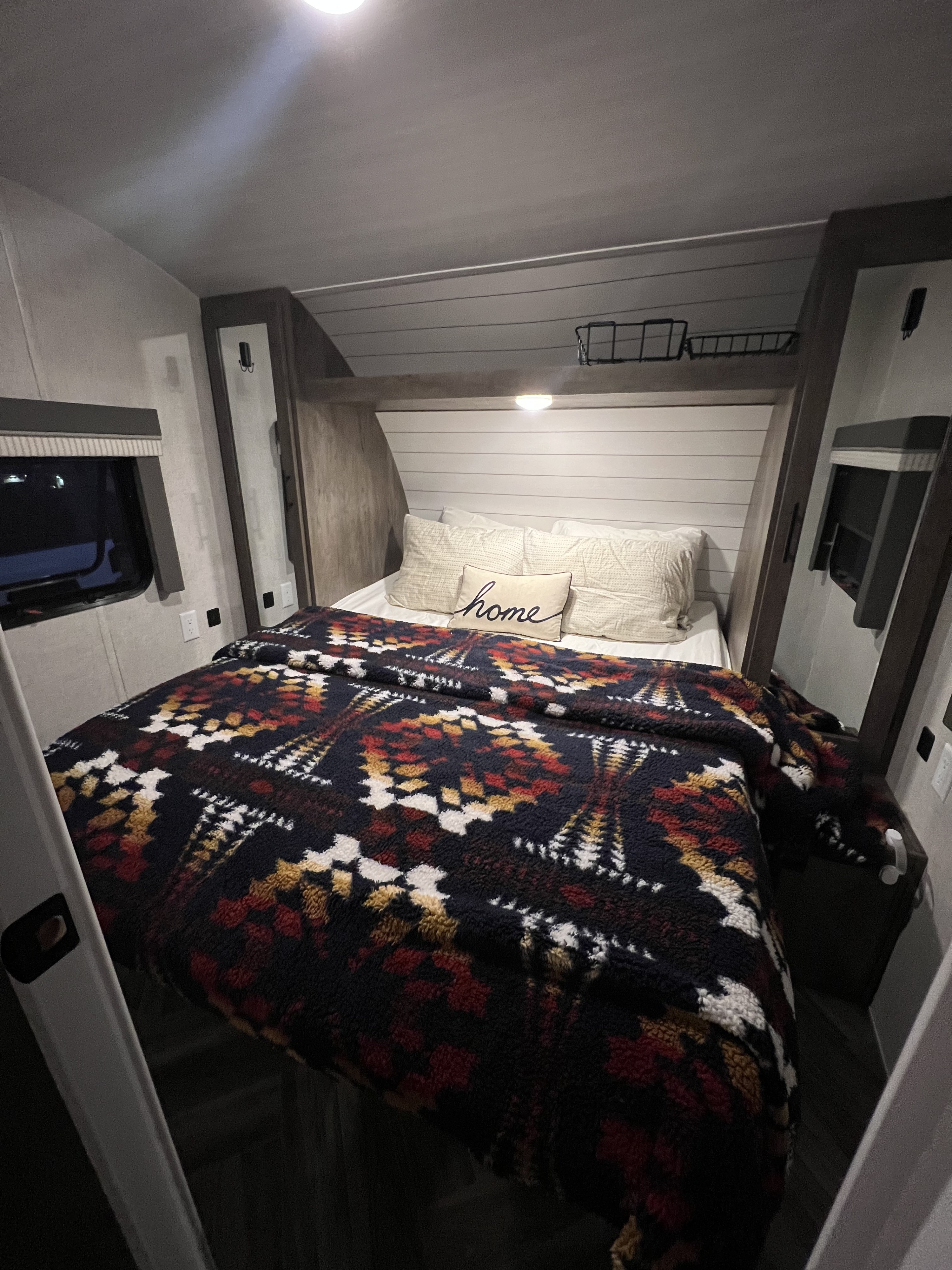 Inside a comfy RV rental bedroom: bed with vibrant patterned blanket, "home" pillow, storage, and window.. Forest River Wildwood X-Lite 2022