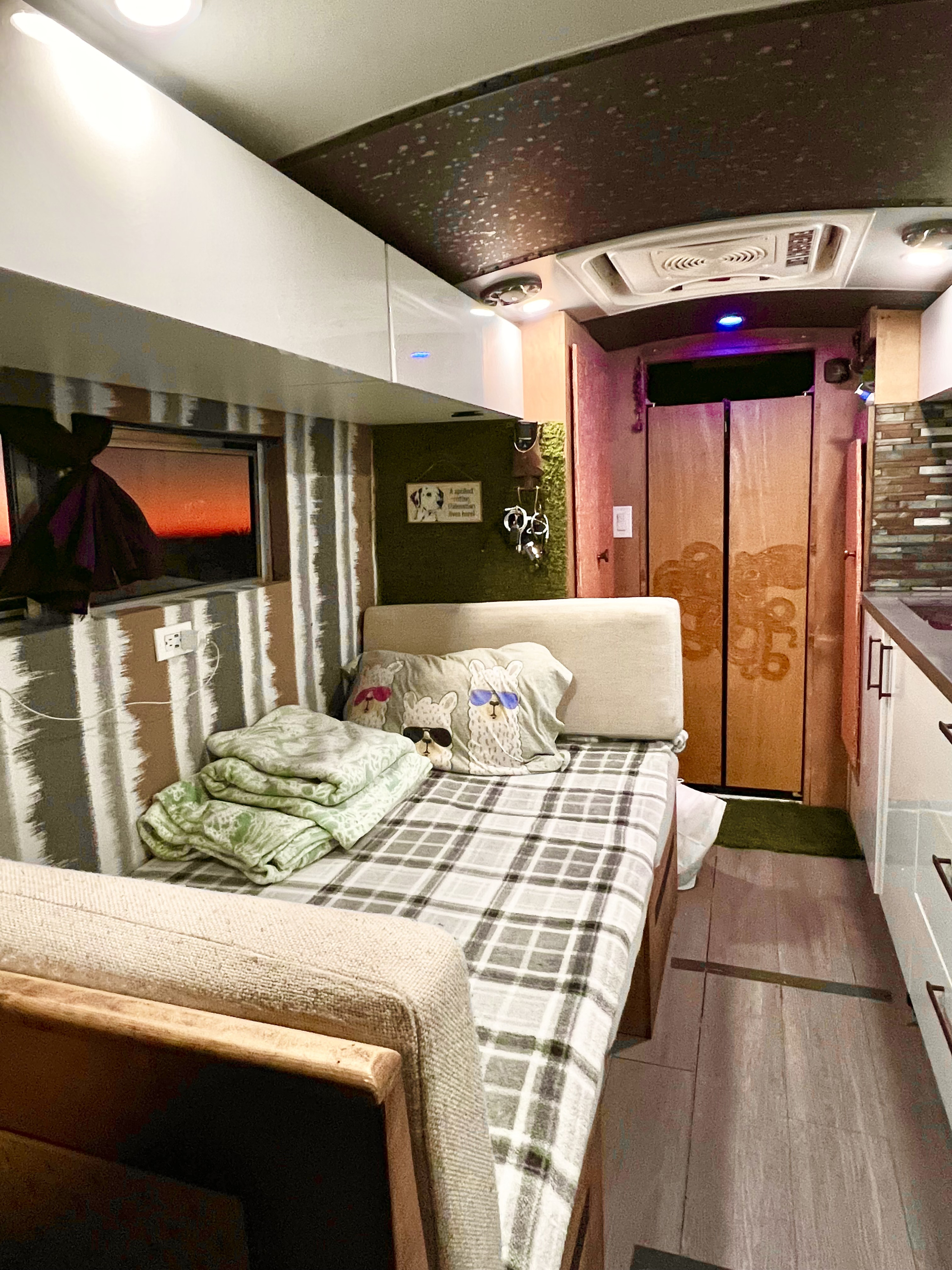 Explore this cozy RV rental interior, featuring a bed with fun llama pillows, compact kitchen, and sunset window.. Chevrolet Off Grid Skoolie 2012