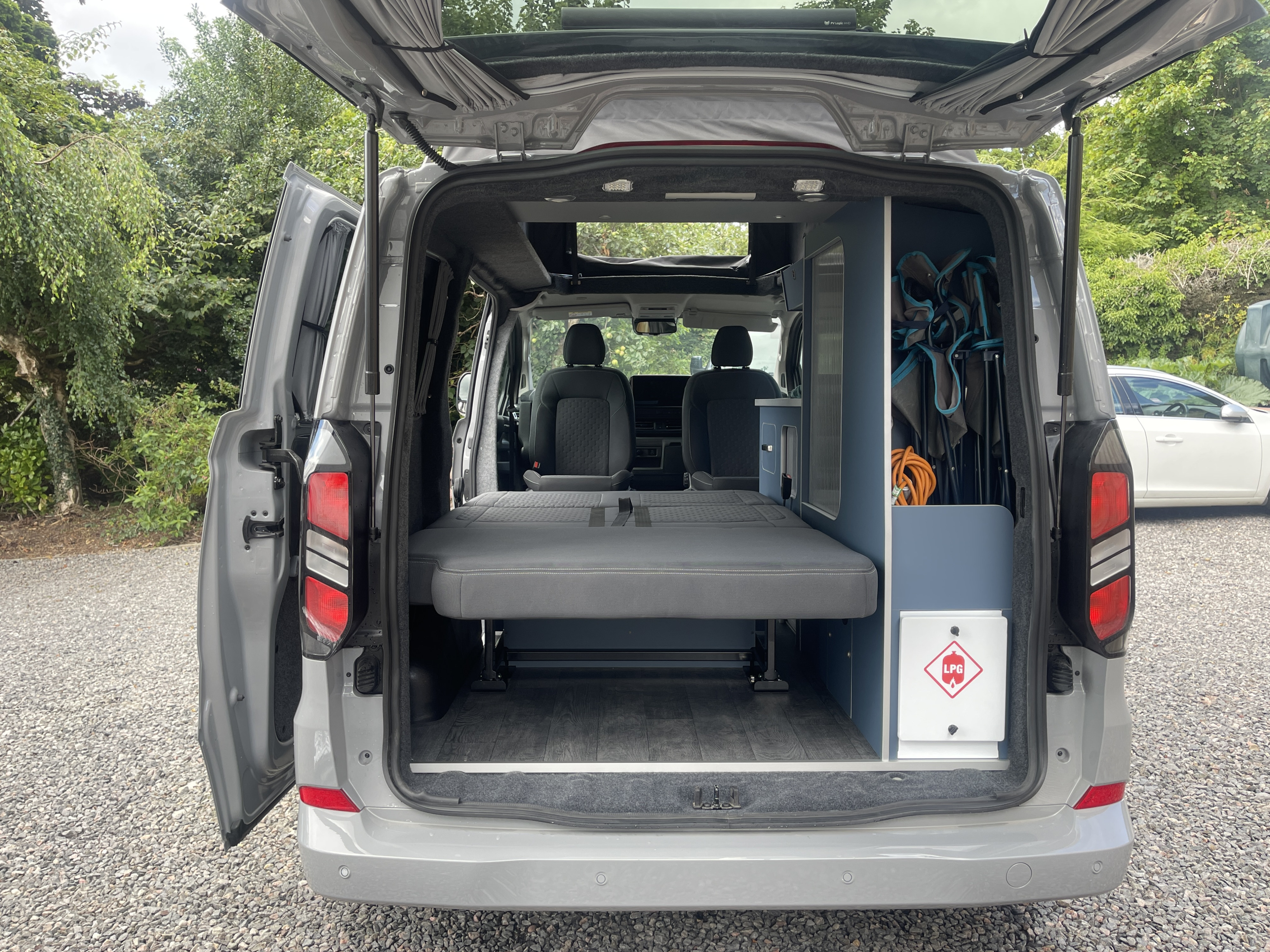 RV rental campervan interior, seen from the rear with open doors. Features a convertible bed, built-in storage, and camping gear.. Ford Transit Custom 2+2 berth Pop Top, Automatic 2025