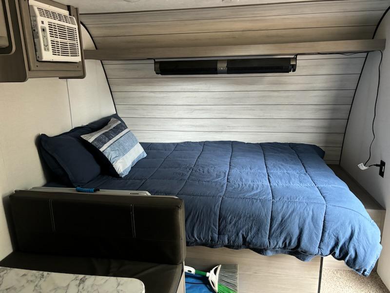 An RV rental's compact interior featuring a made bed with blue linens, wall-mounted AC, and built-in dinette.. Dutchmen Lantern 17b 2024