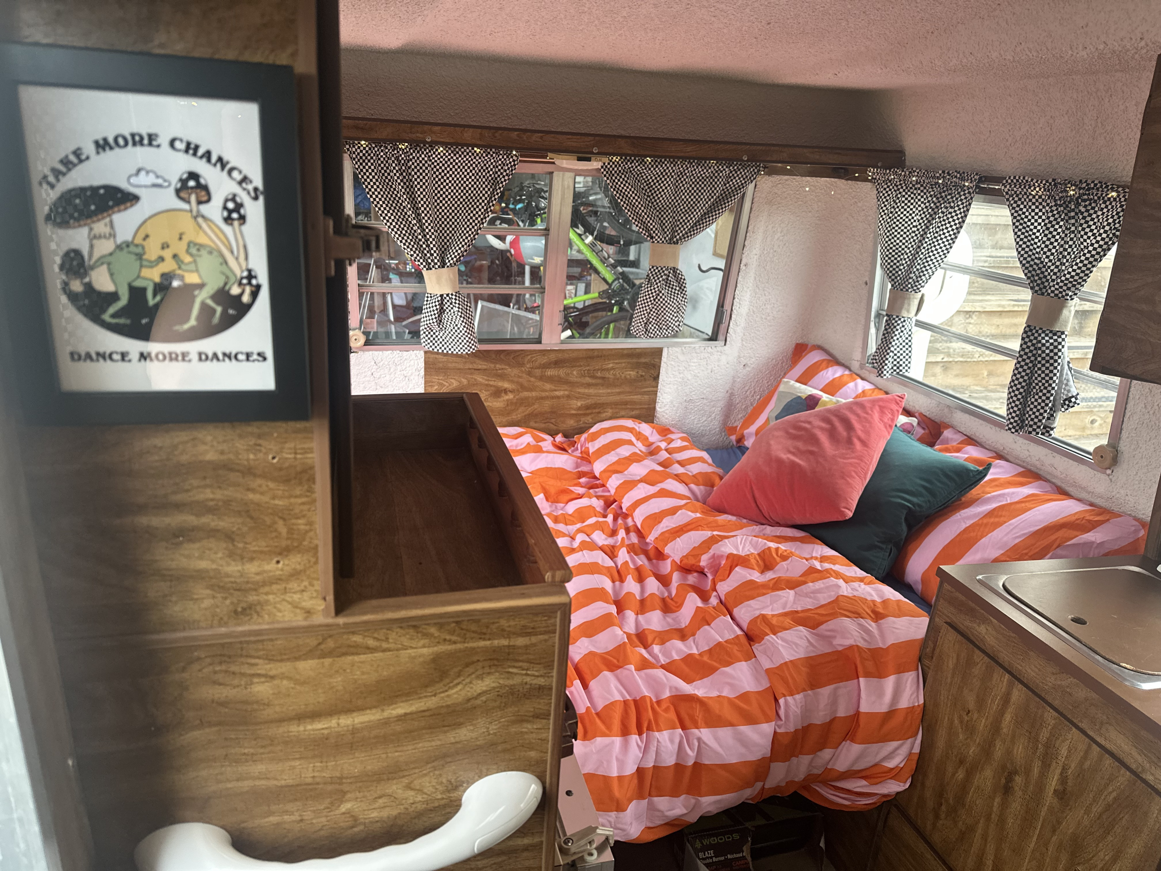 An inviting RV rental interior featuring a striped bed, checkered curtains, unique art, and a small kitchen sink.. Trillium Rv Jubilee 1980