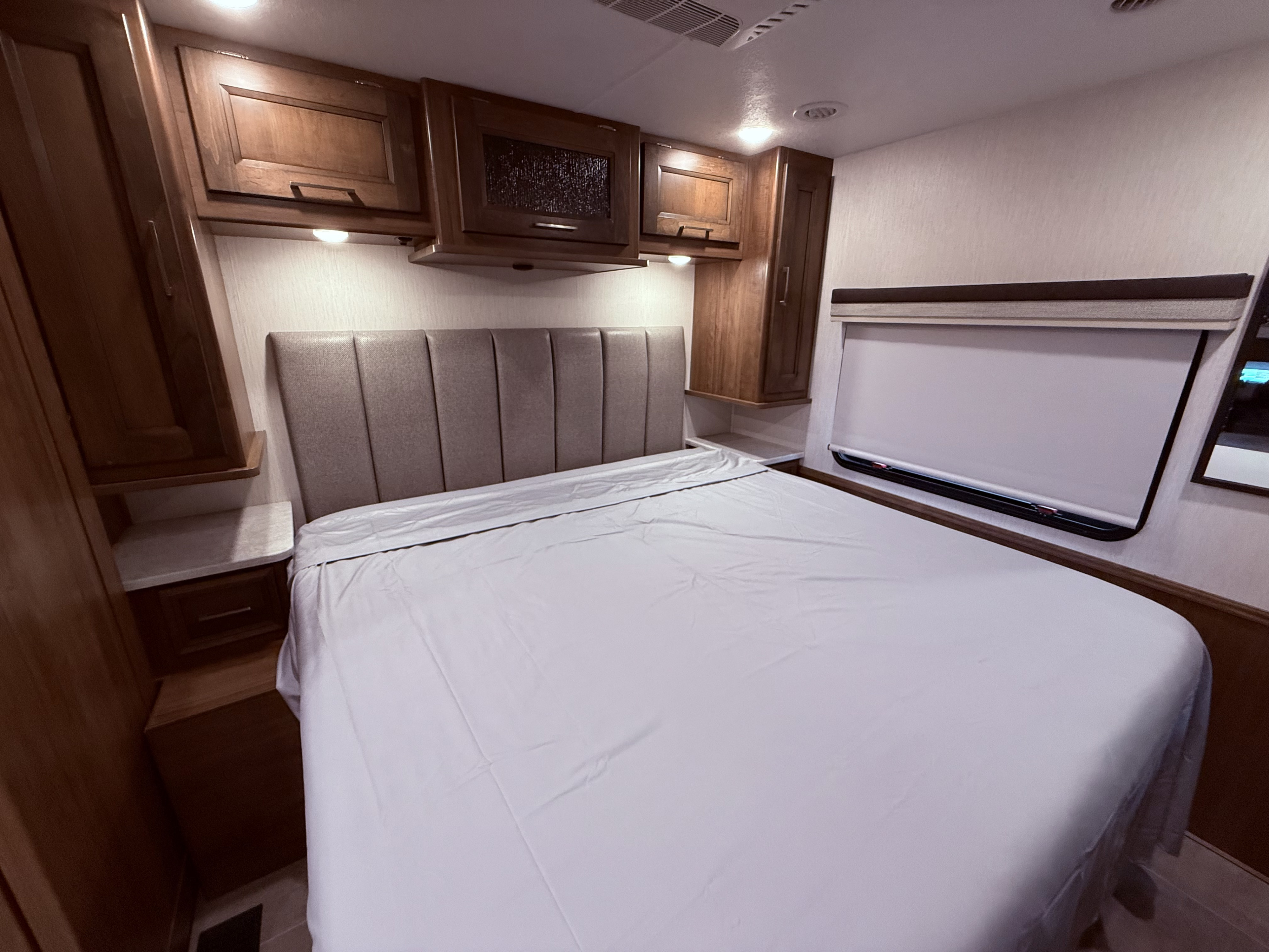 Interior of an RV rental bedroom with a large bed, padded headboard, wooden cabinets, and a window.. Coachman Encore 2021