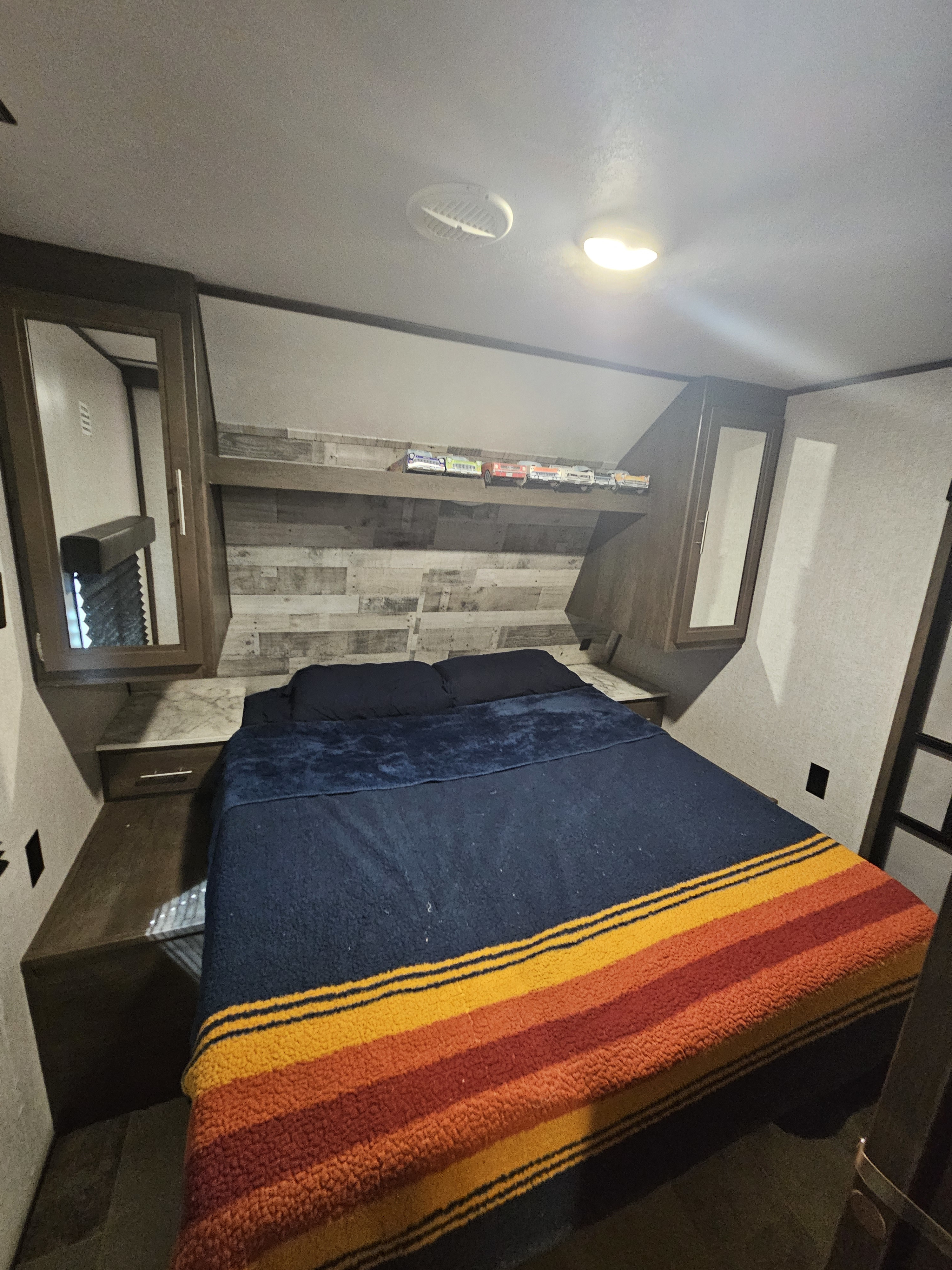 RV rental bedroom showcasing a comfy bed with a striped blanket, integrated storage, and wood-paneled headboard. Ideal for compact travel.. Dutchmen Coleman Lantern 2021