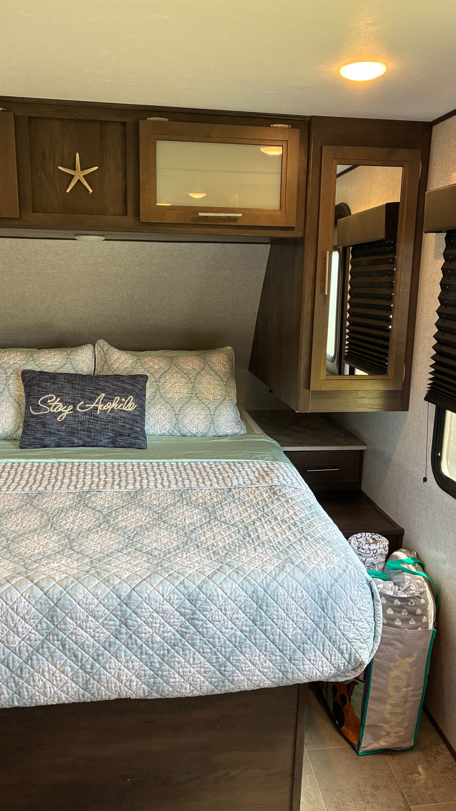 Cozy RV rental bedroom with a neatly made bed, patterned quilt, "Stay Awhile" pillow, and overhead cabinets.. Dutchmen Coleman Lantern LT 2022