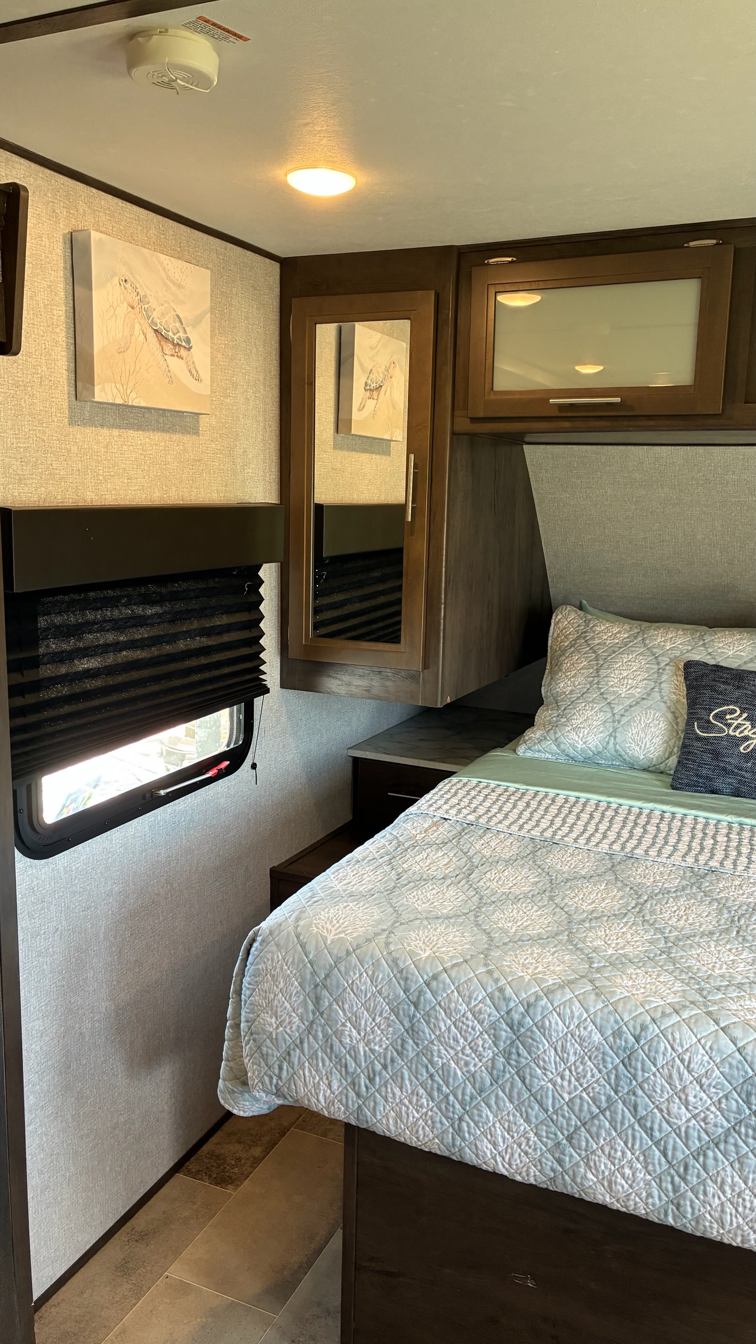 Cozy RV rental bedroom with light blue coral-patterned bedding, a compact window with black blinds, and turtle wall art.. Dutchmen Coleman Lantern LT 2022