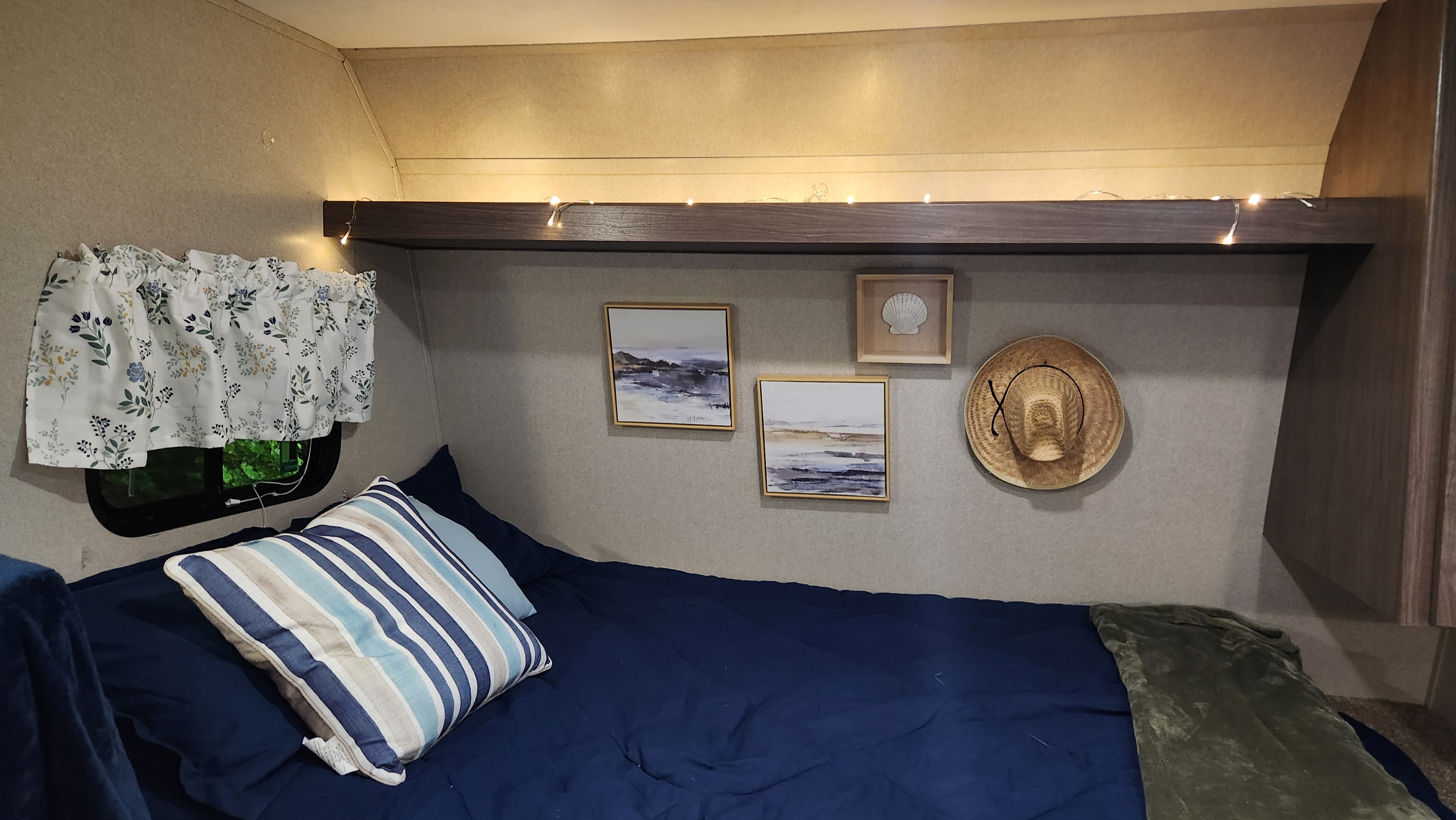 Cozy RV rental bedroom featuring navy blue bedding, a striped pillow, string lights, & framed art with a straw hat.. Keystone RV Hideout LHS 2019