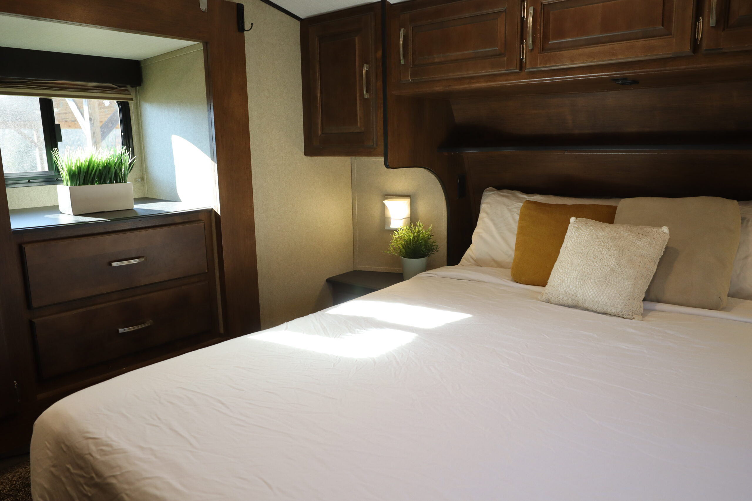 Step inside this cozy RV rental bedroom featuring a comfy bed, dark wood cabinets, and a bright window with plants.. Grand Design Reflection 2019