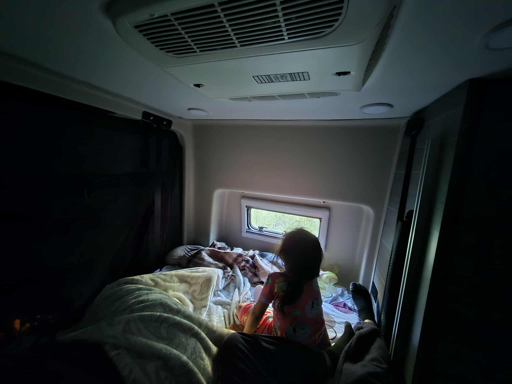 Inside a cozy RV rental: A child sits on the bed looking out the window at trees. Comfort for family travel.. Entegra Launch Sprinter High Roof AWD 4x4 Camper Van 2025