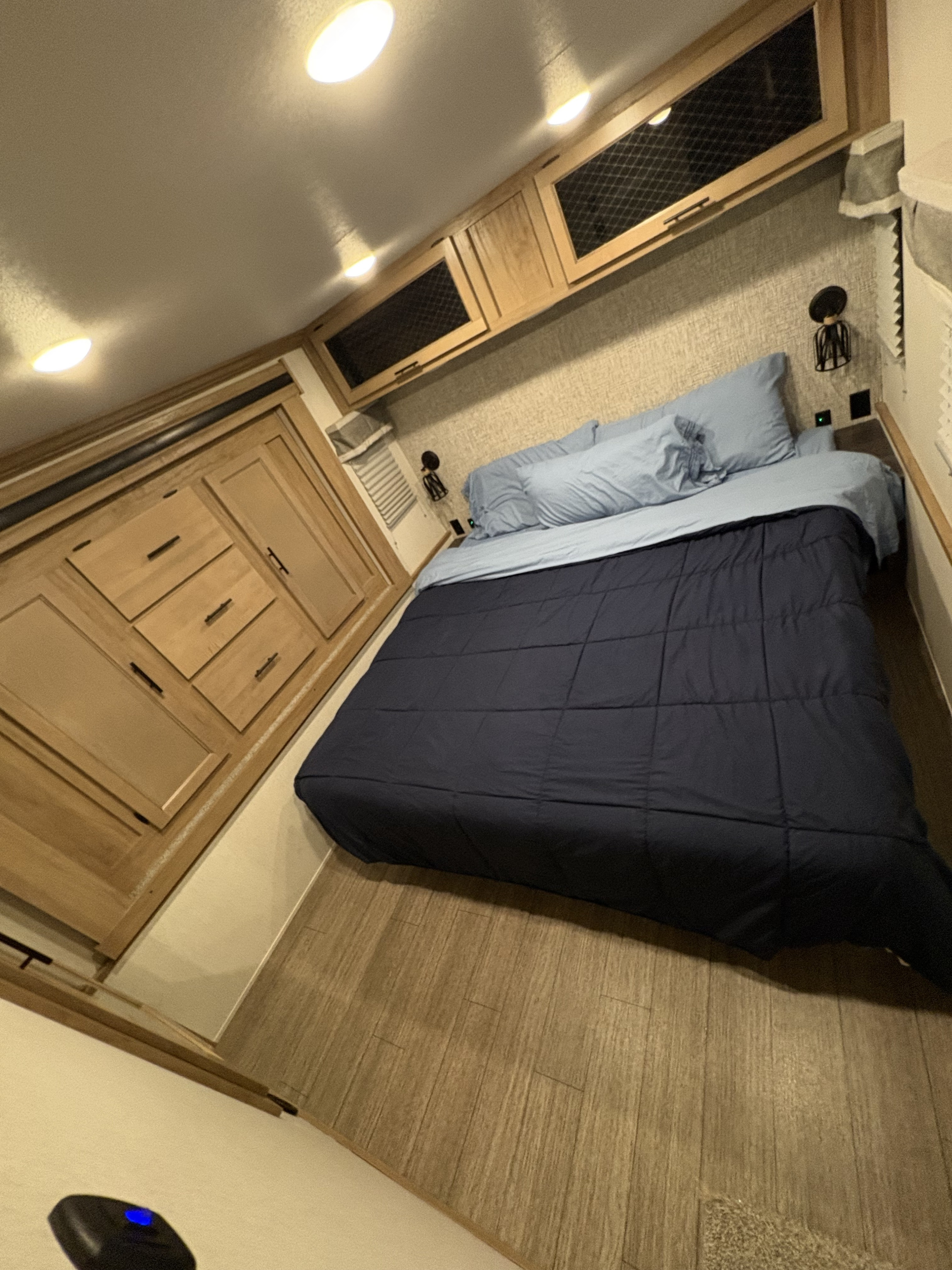 Cozy RV rental bedroom featuring a bed with blue bedding, wooden cabinets, and ceiling lights, ideal for compact travel.. Forest River Sabre 2023