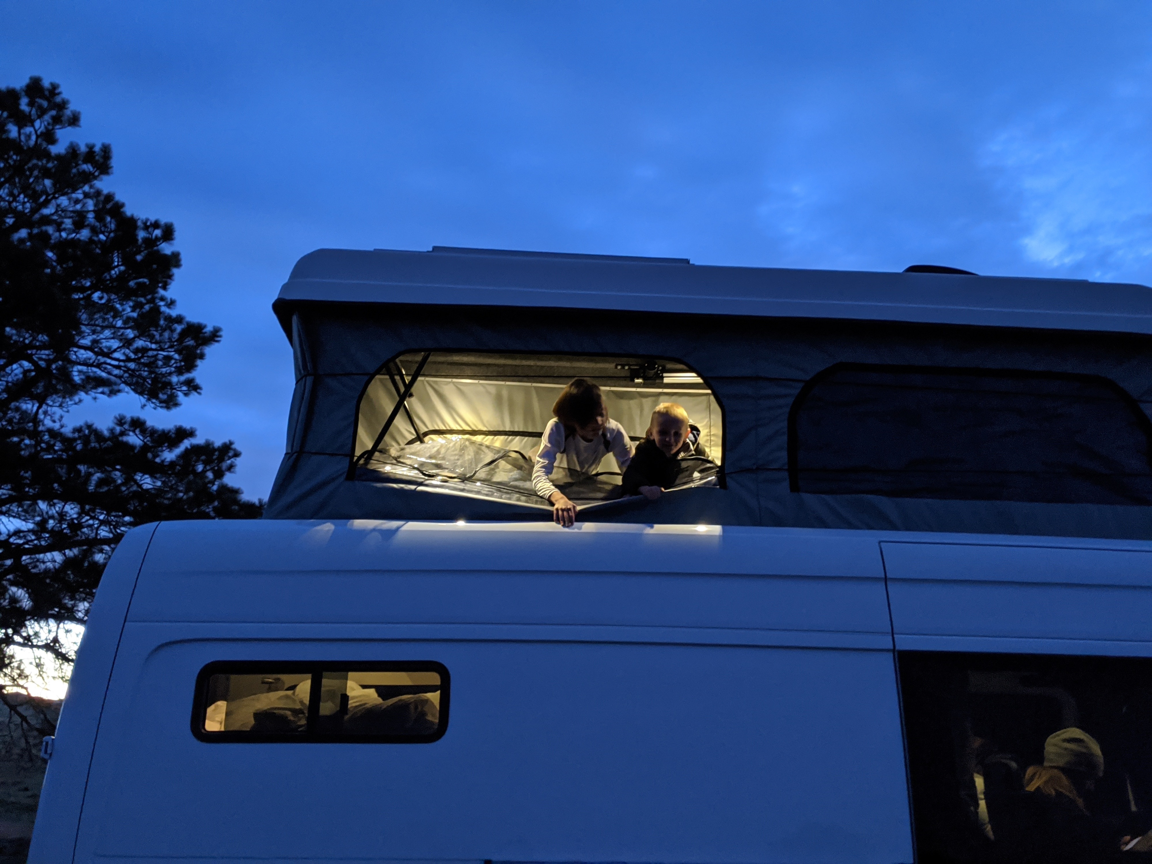 Two kids peer from a lit pop-top camper, an **RV rental** perfect for evening adventures.. Mercedes-Benz Sprinter 2020