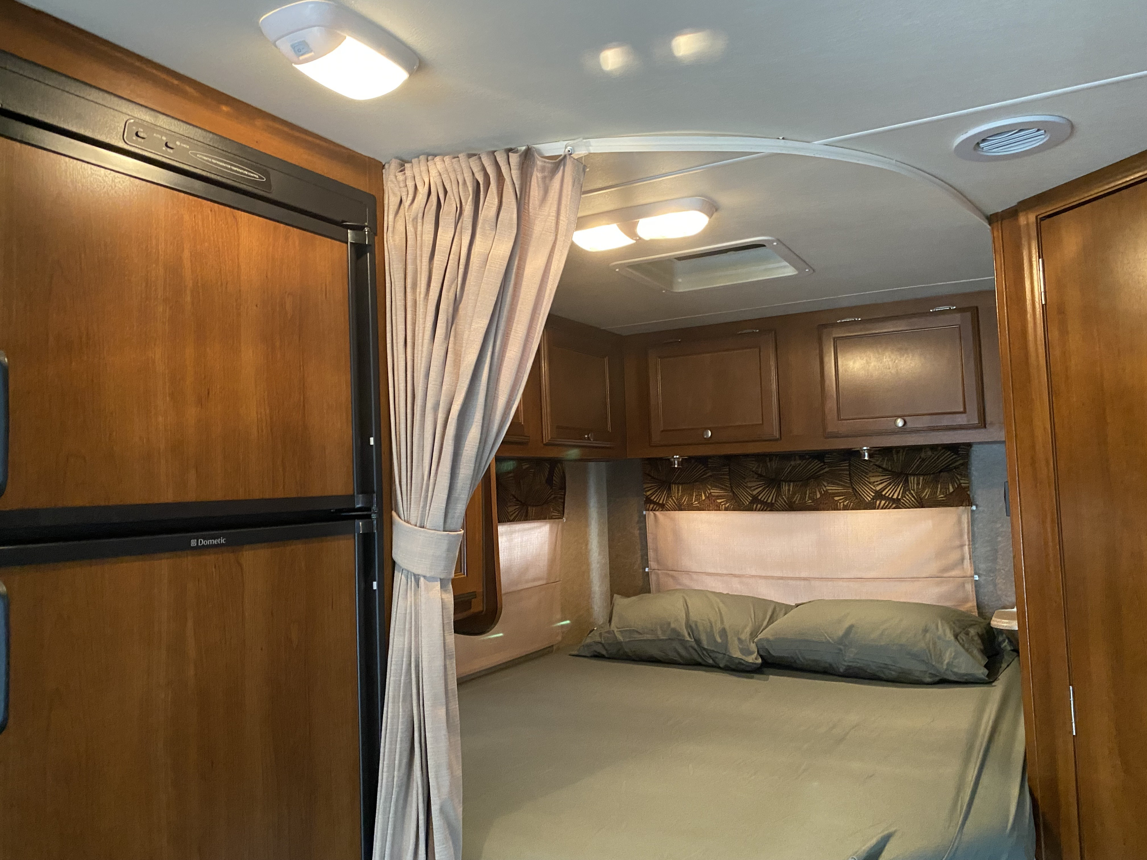 RV rental bedroom with a comfortable bed, green sheets, and overhead cabinets. Features a Dometic fridge and privacy curtain.. Forest River Coachmen Leprechaun 2015