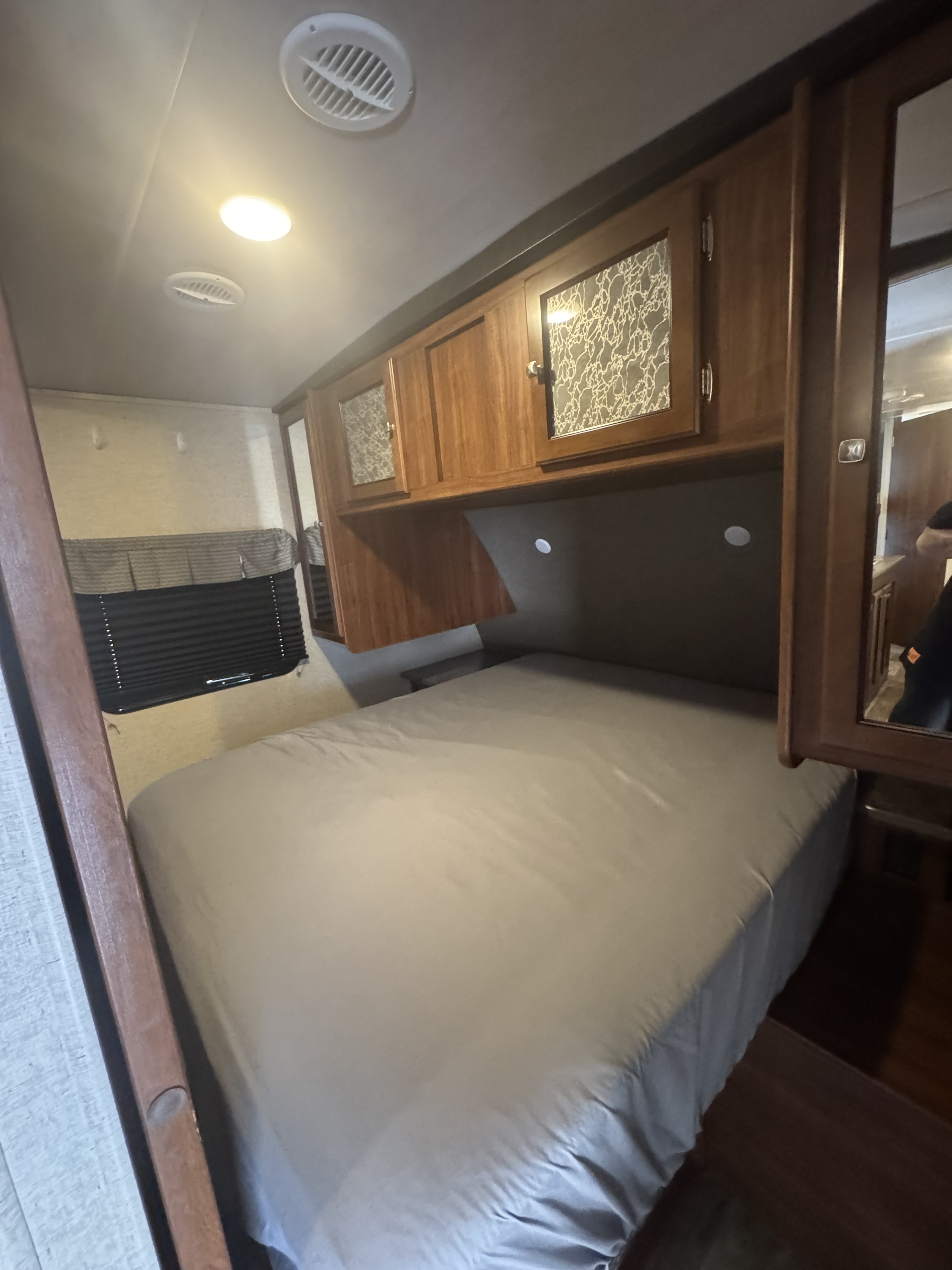 RV rental bedroom with a comfy bed, overhead wooden cabinets, and window blinds. Bright ceiling lights illuminate the space.. Heartland RVs Wilderness 2020
