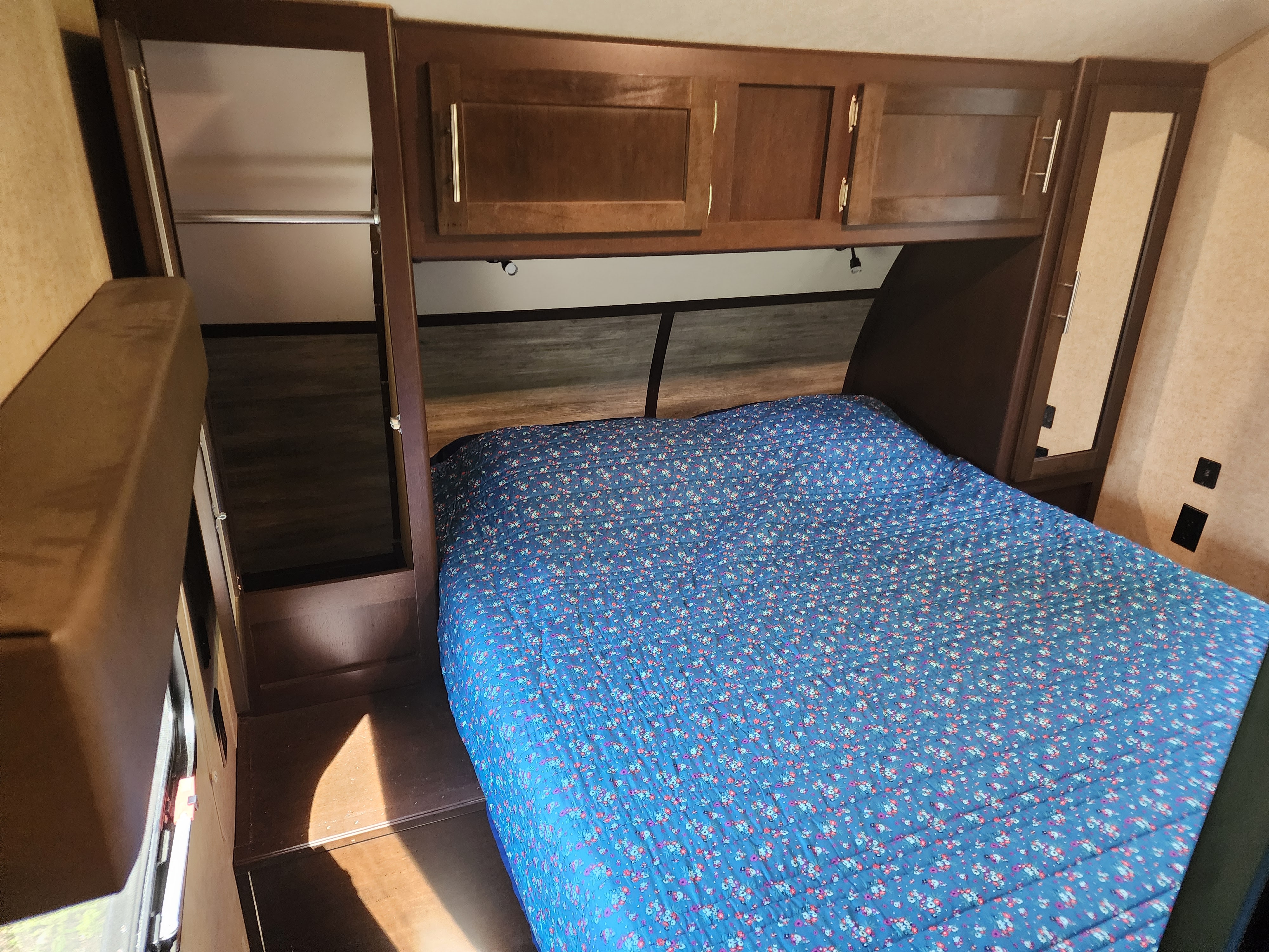 Cozy RV rental bedroom featuring a bed with a blue floral comforter, dark wood cabinets, and a mirrored closet door.. Forest River Cherokee 2018