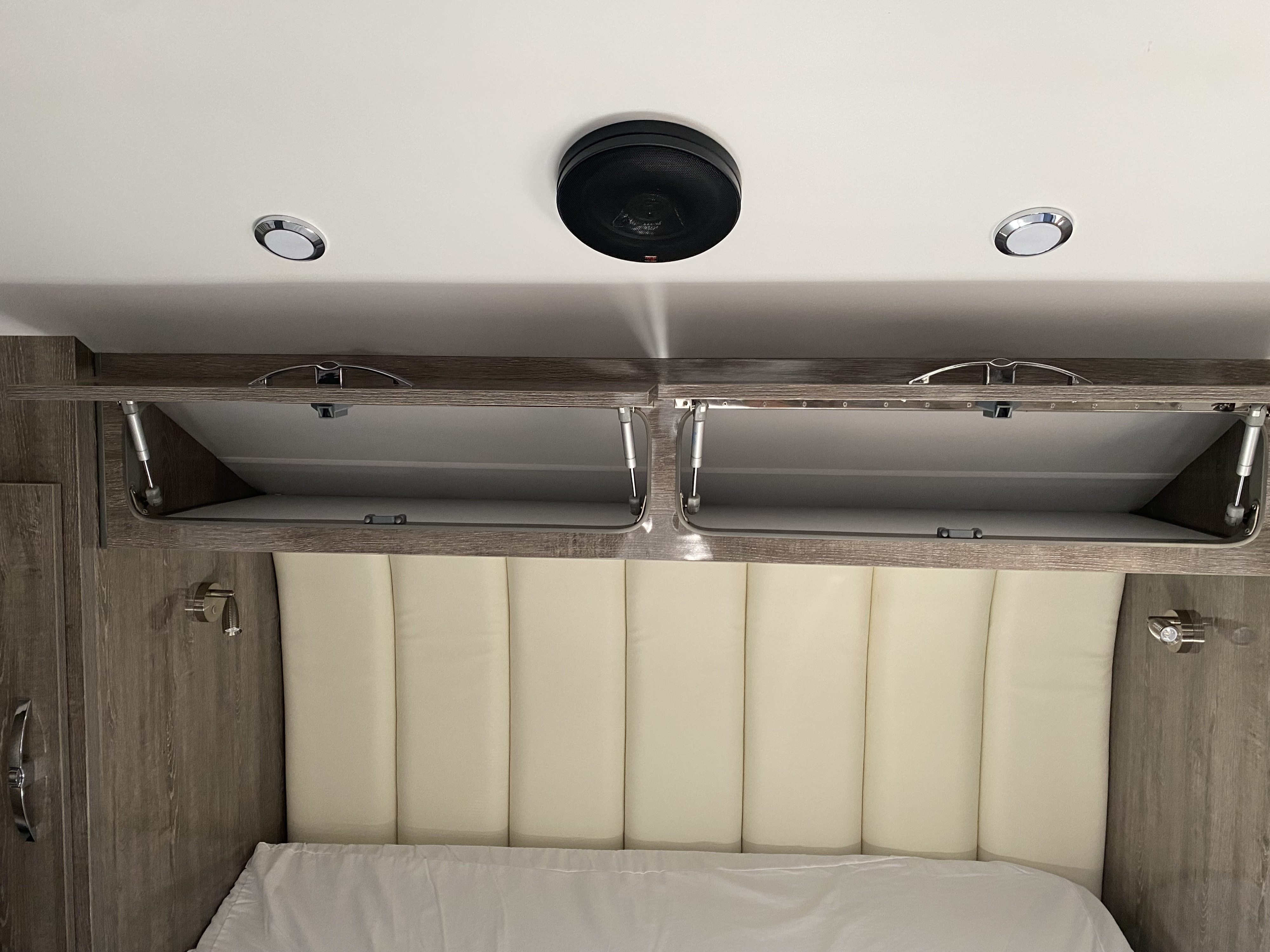 Comfortable RV rental bedroom with padded headboard, open overhead storage cabinets, and ceiling-mounted speaker/lights.. Black Series Black Series Trailer 2021