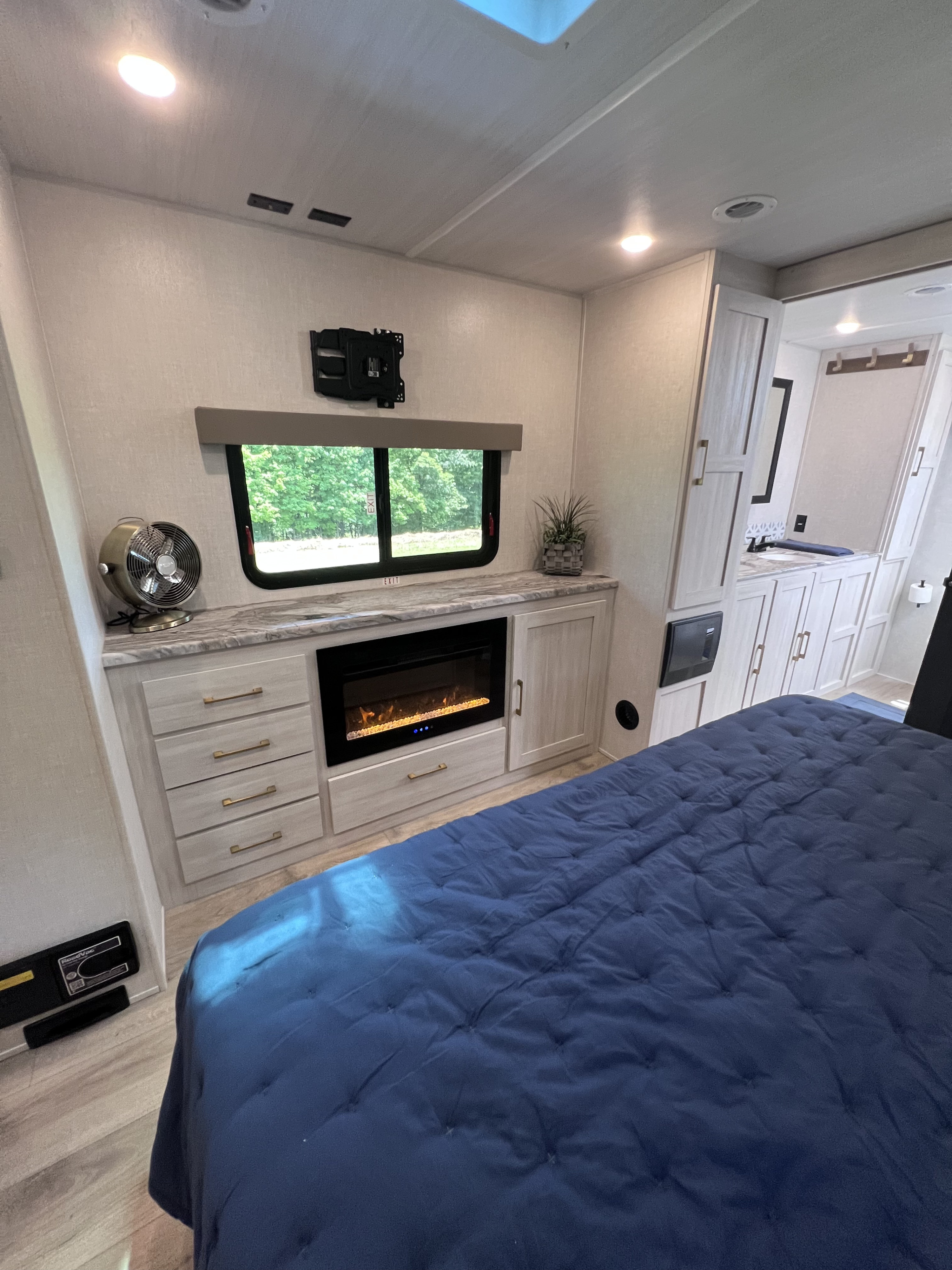 Modern RV rental bedroom featuring a plush blue bed, electric fireplace, and large window with nature views.. Forest River Grand Surveyor 2024