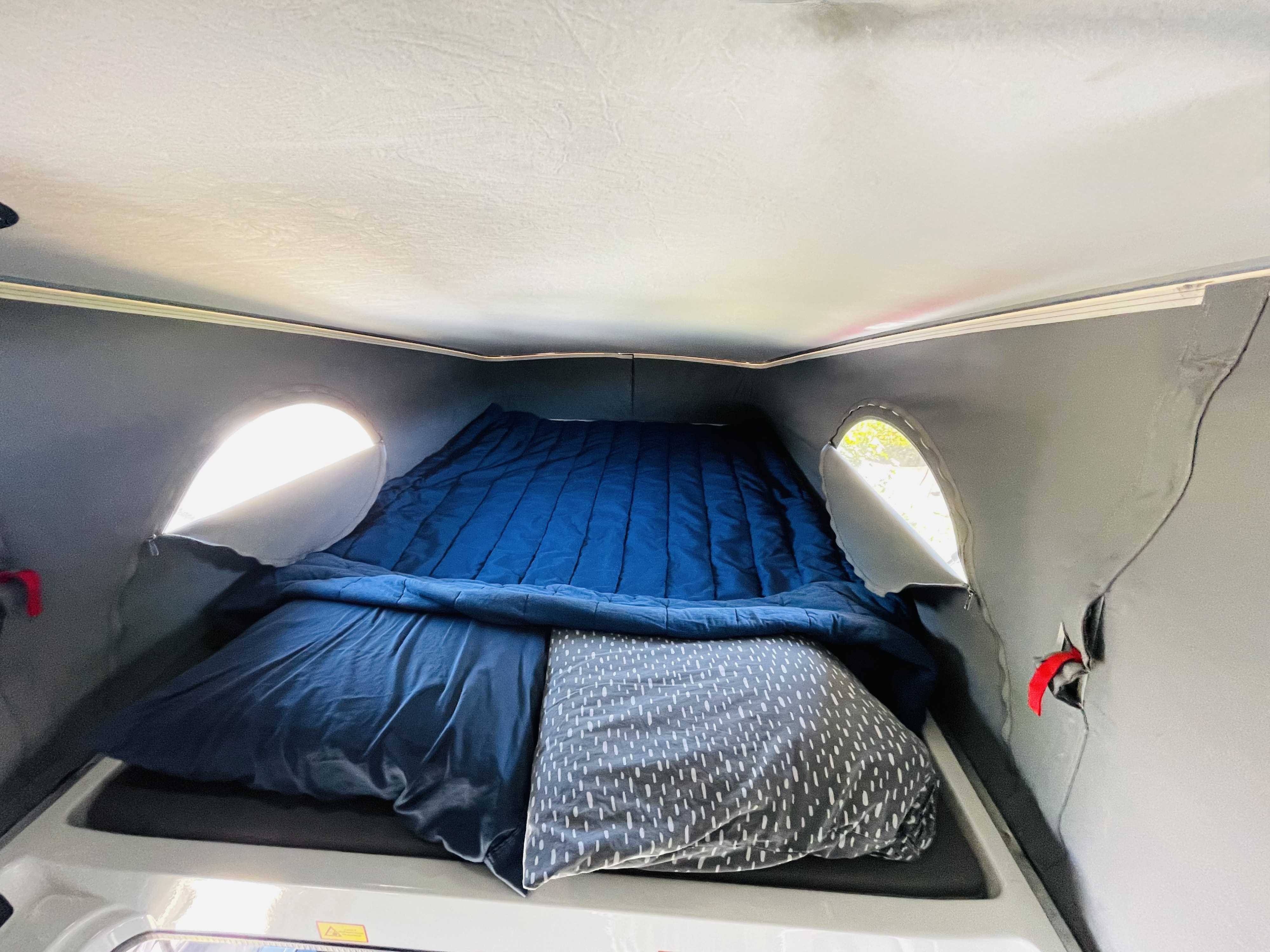 RV rental pop-top sleeping area with a blue bed and grey patterned pillow. Bright light streams through unique round, zipped windows.. Winnebago Solis 2022