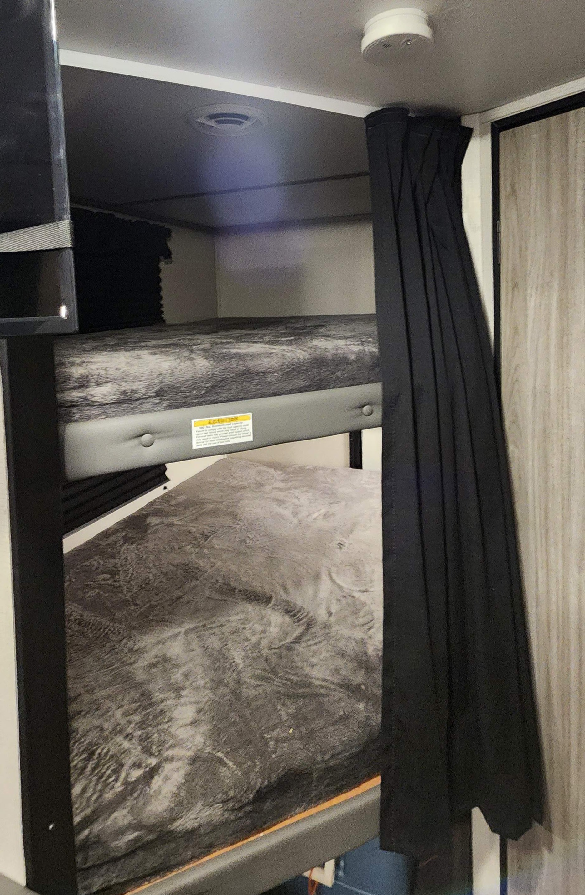 Inside an RV rental: two gray-patterned bunk beds with a privacy curtain, overhead vent, and smoke detector.. Keystone RV Passport SL 2022