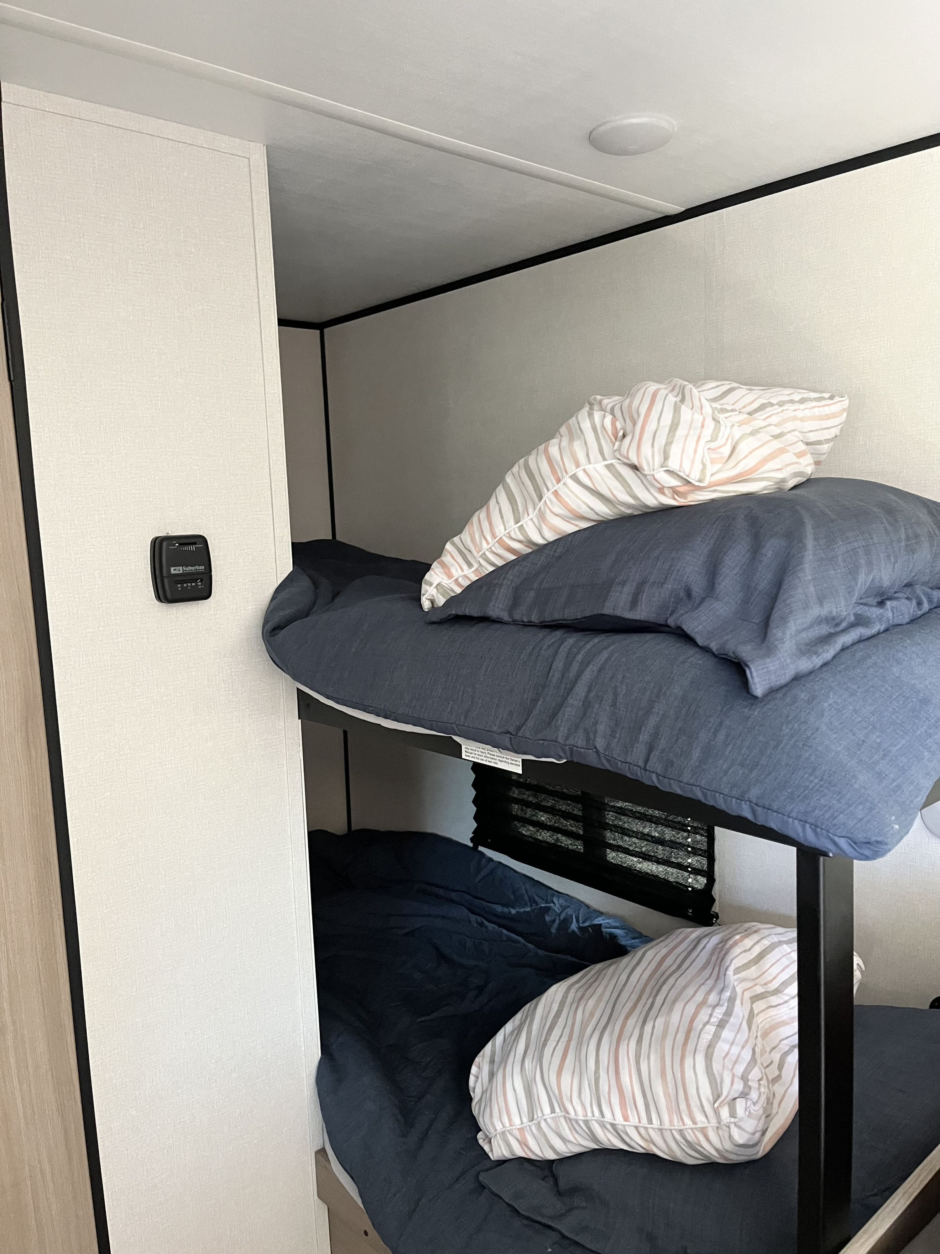 Inside an RV rental, showing compact bunk beds with blue bedding, a striped pillow, and a wall thermostat.. Coleman 7B 2025
