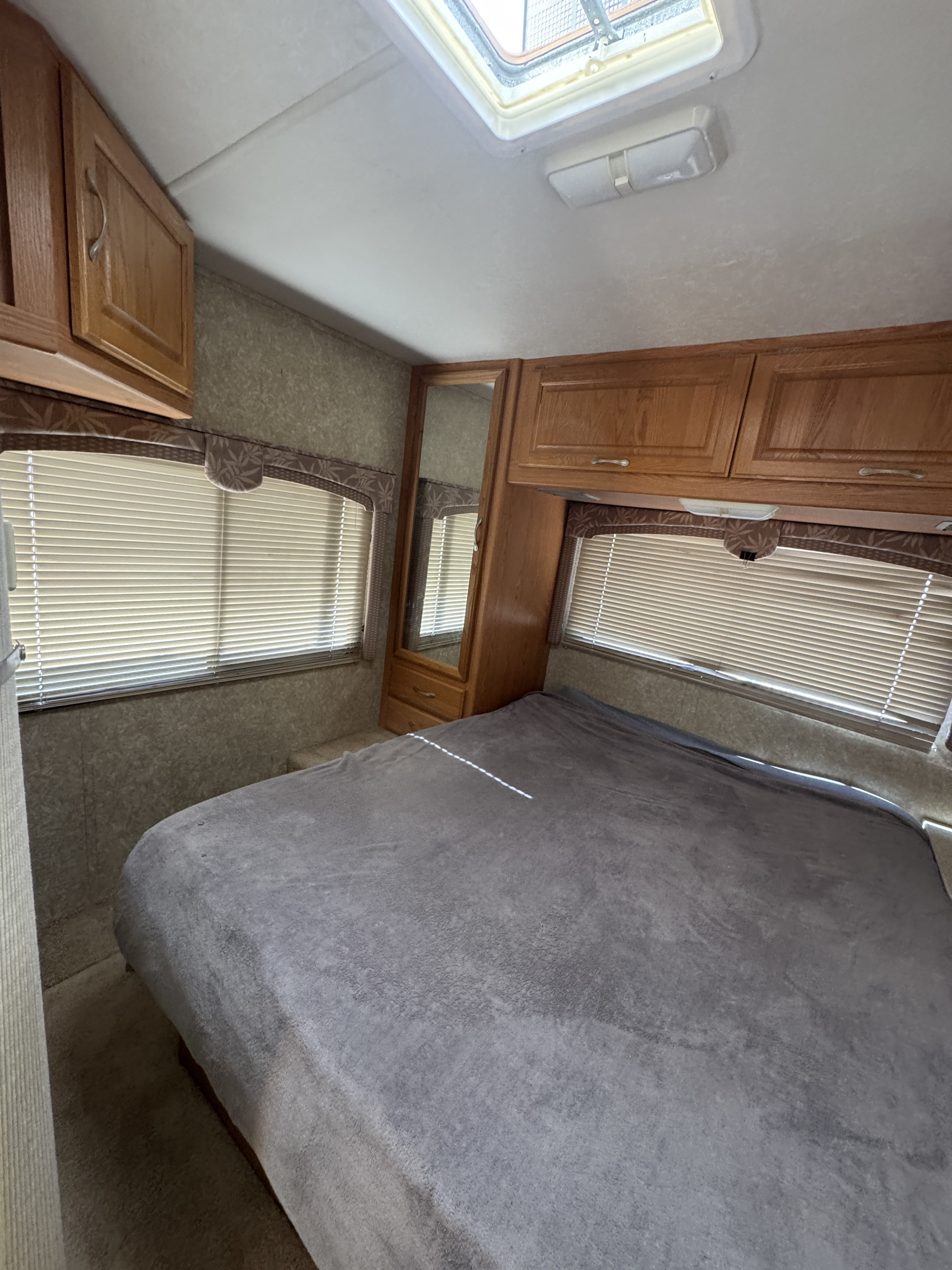Cozy RV rental bedroom interior featuring a comfortable bed, wooden cabinets, multiple windows with blinds, and a ceiling skylight.. Dutchmen Express 2005