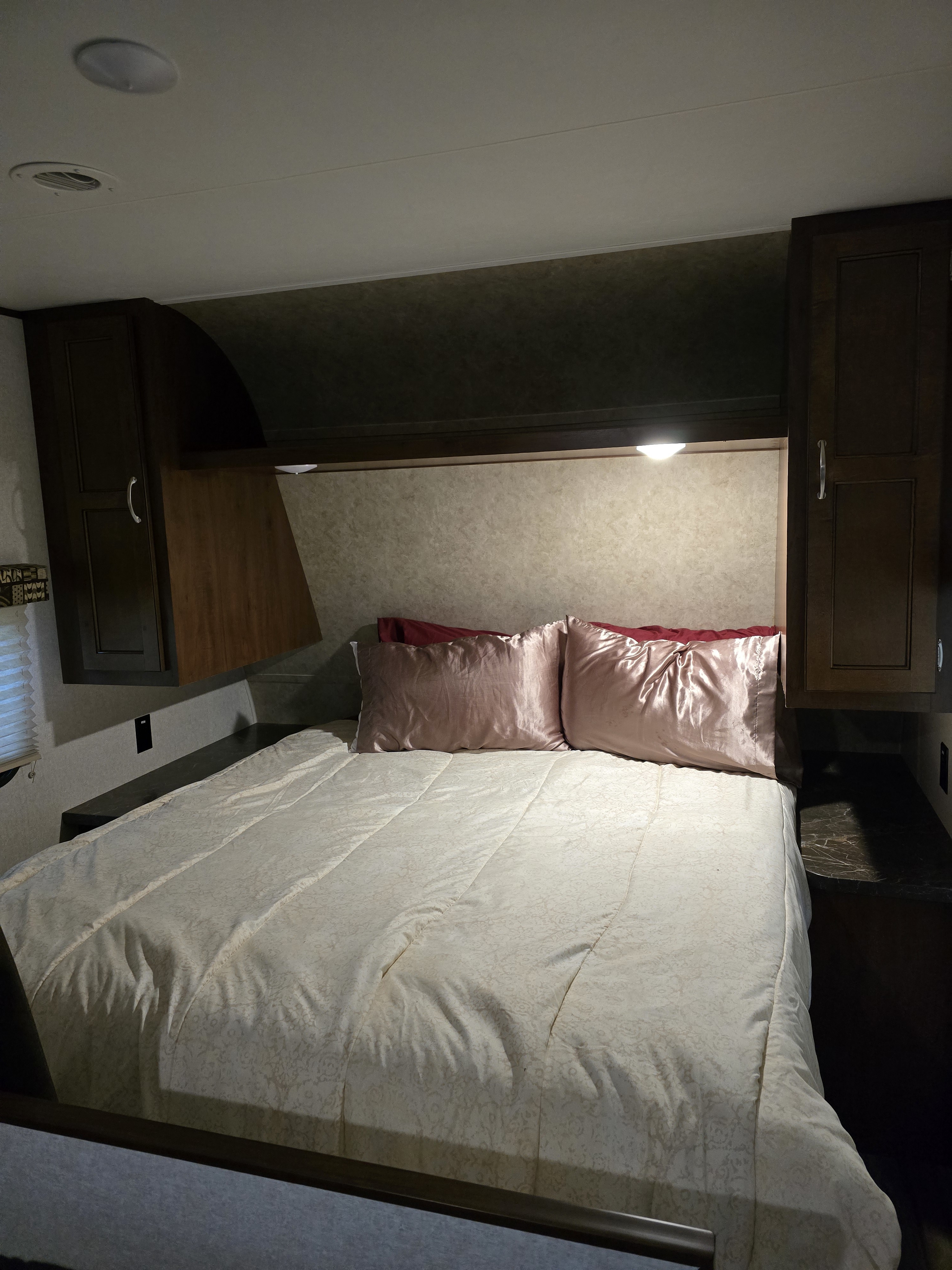A compact RV rental bedroom features a neatly made bed with pink pillows, dark wood cabinets, and ceiling lights, ideal for travel.. Jayco Jay Flight SLX 2017