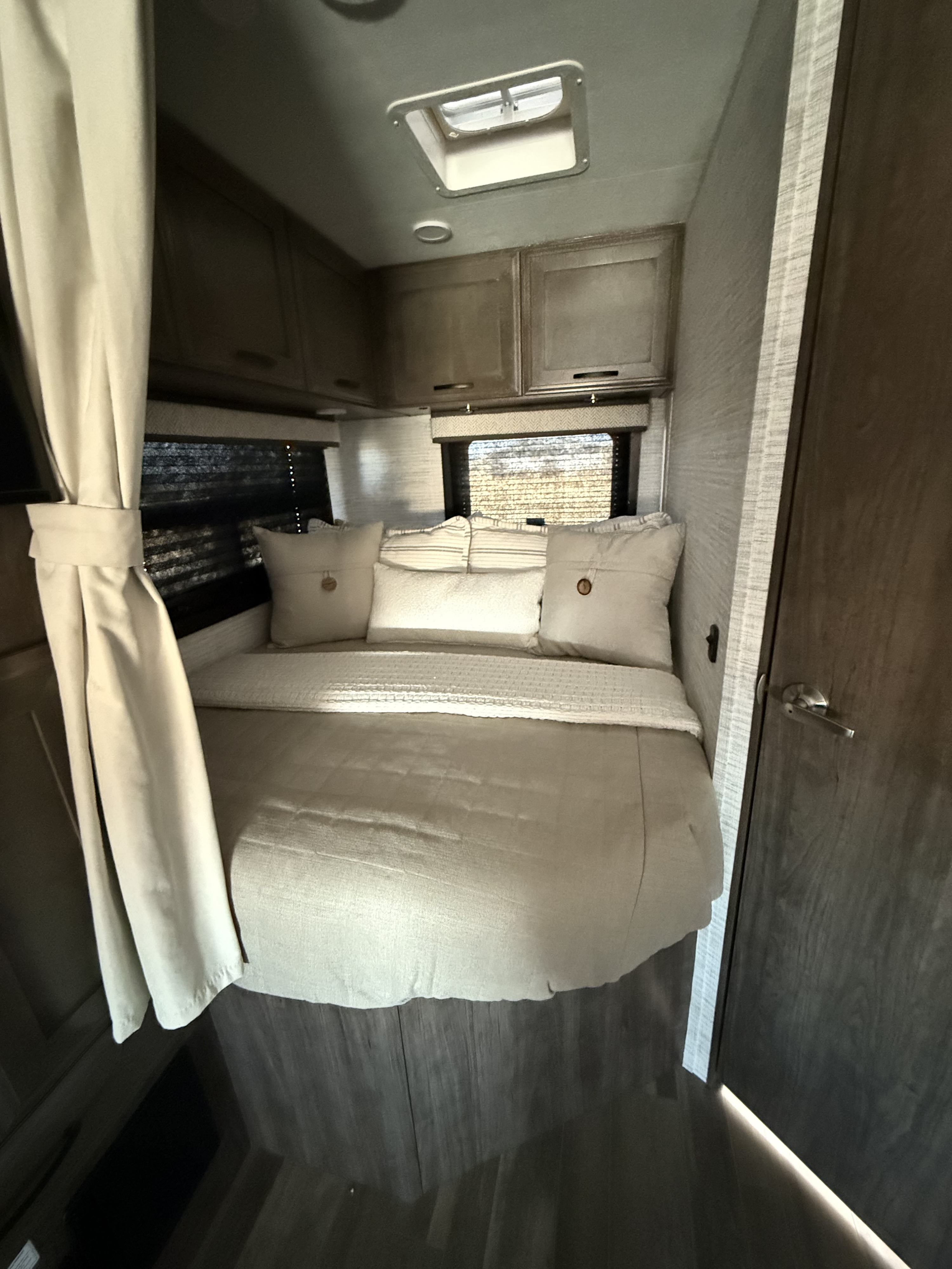 Cozy RV rental bedroom with bed, cabinets, and skylight. Light curtains and a dark wood door are visible.. Entegra Odyssey 2025
