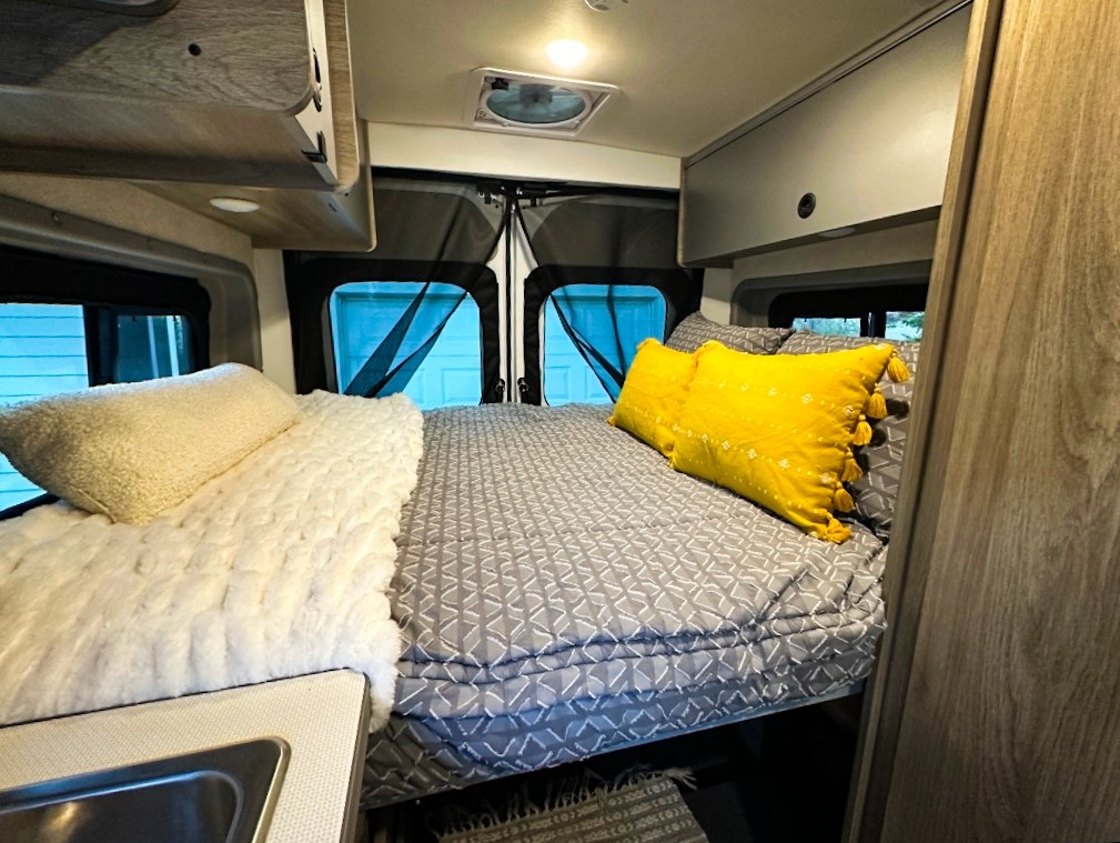 Explore this RV rental's cozy bedroom featuring two beds with different bedding: white fluffy and gray patterned with yellow pillows.. Winnebago Solis 2022