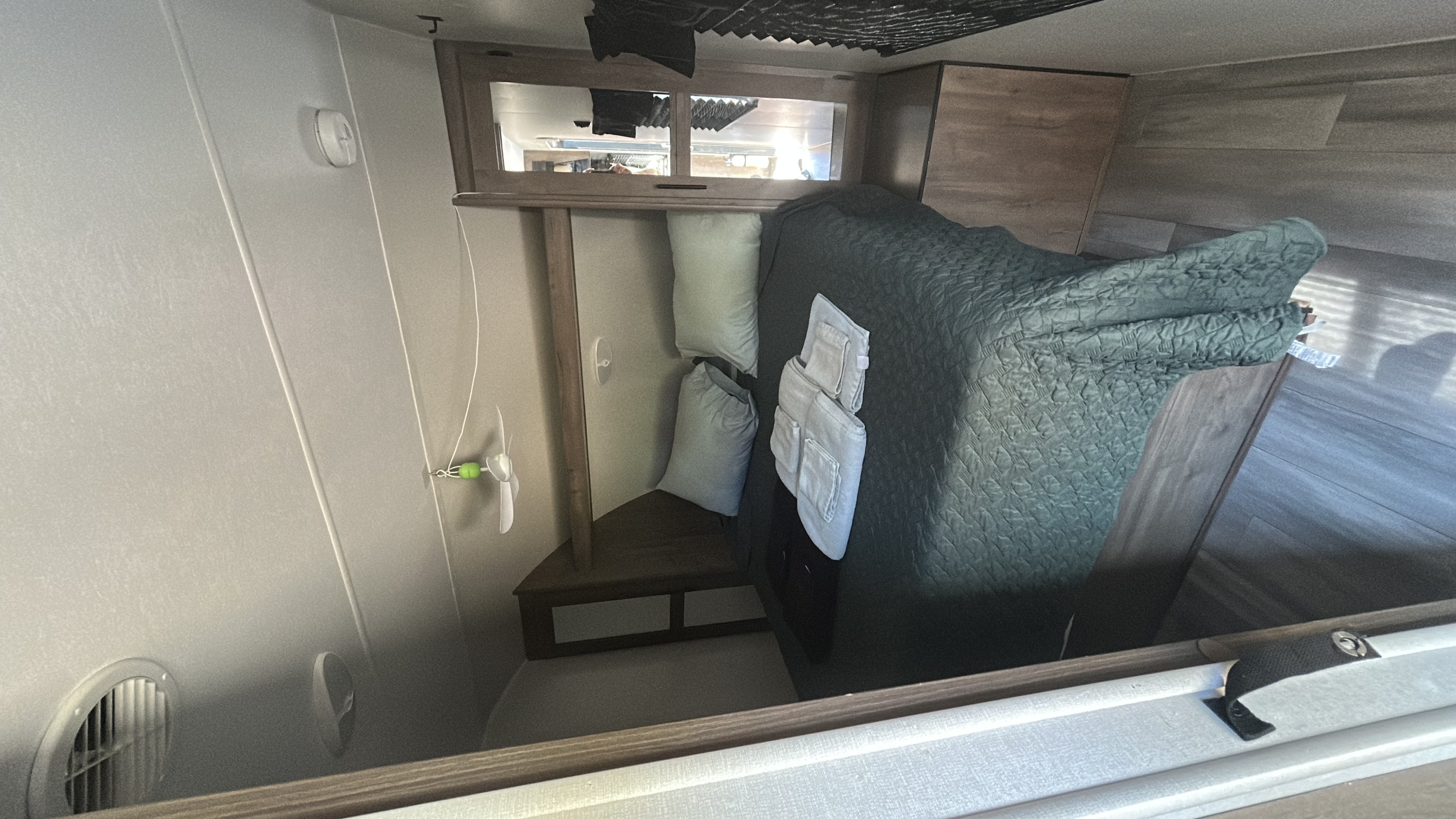 Interior of an RV rental: a cozy bunk with a green quilted comforter, pillows, and a small white fan.. Heartland RVs Pioneer 2024