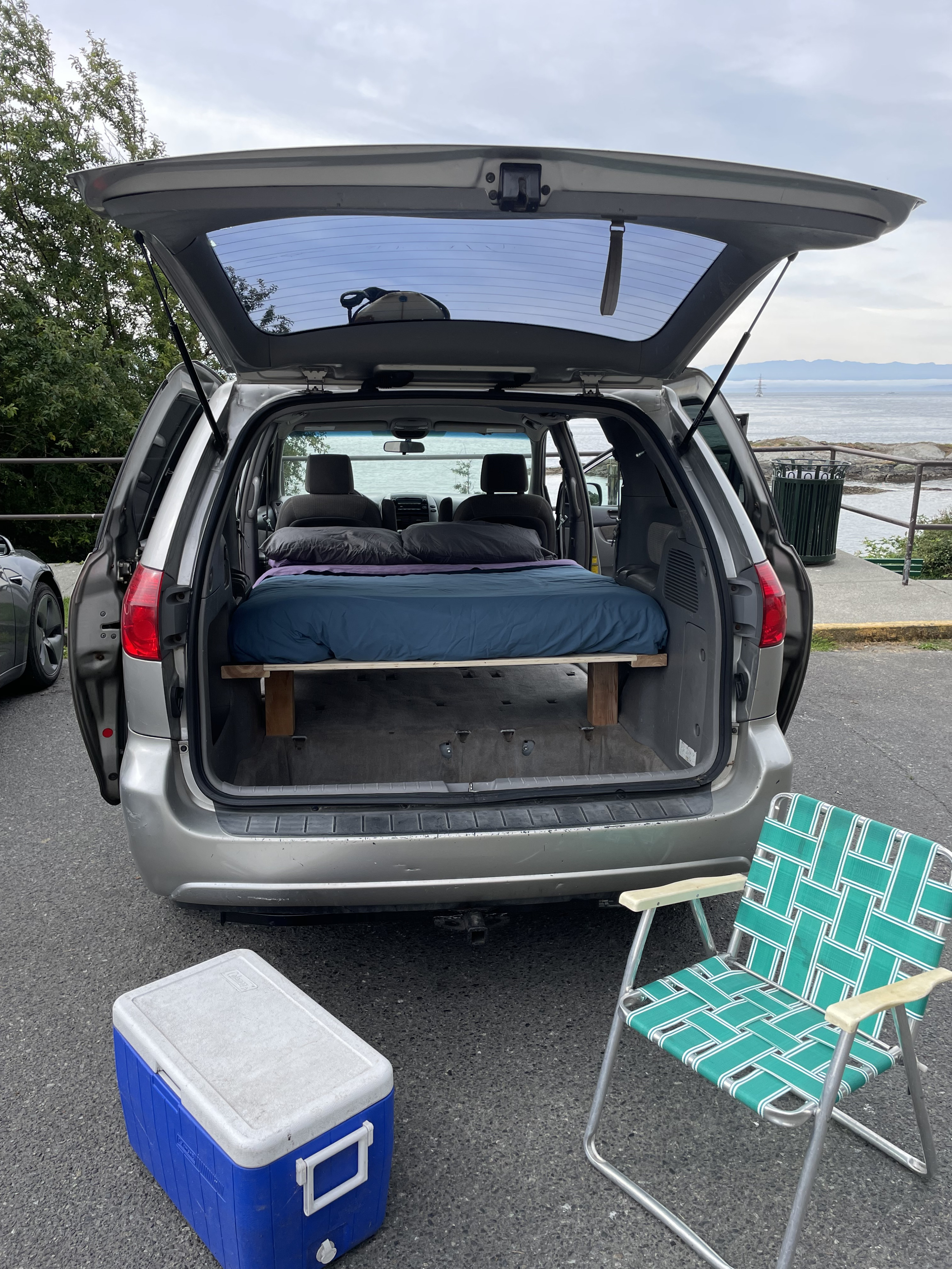 Minivan converted to an RV rental setup with a cozy bed, cooler, and chair, parked by a scenic ocean view.. Toyota Granvia 2008