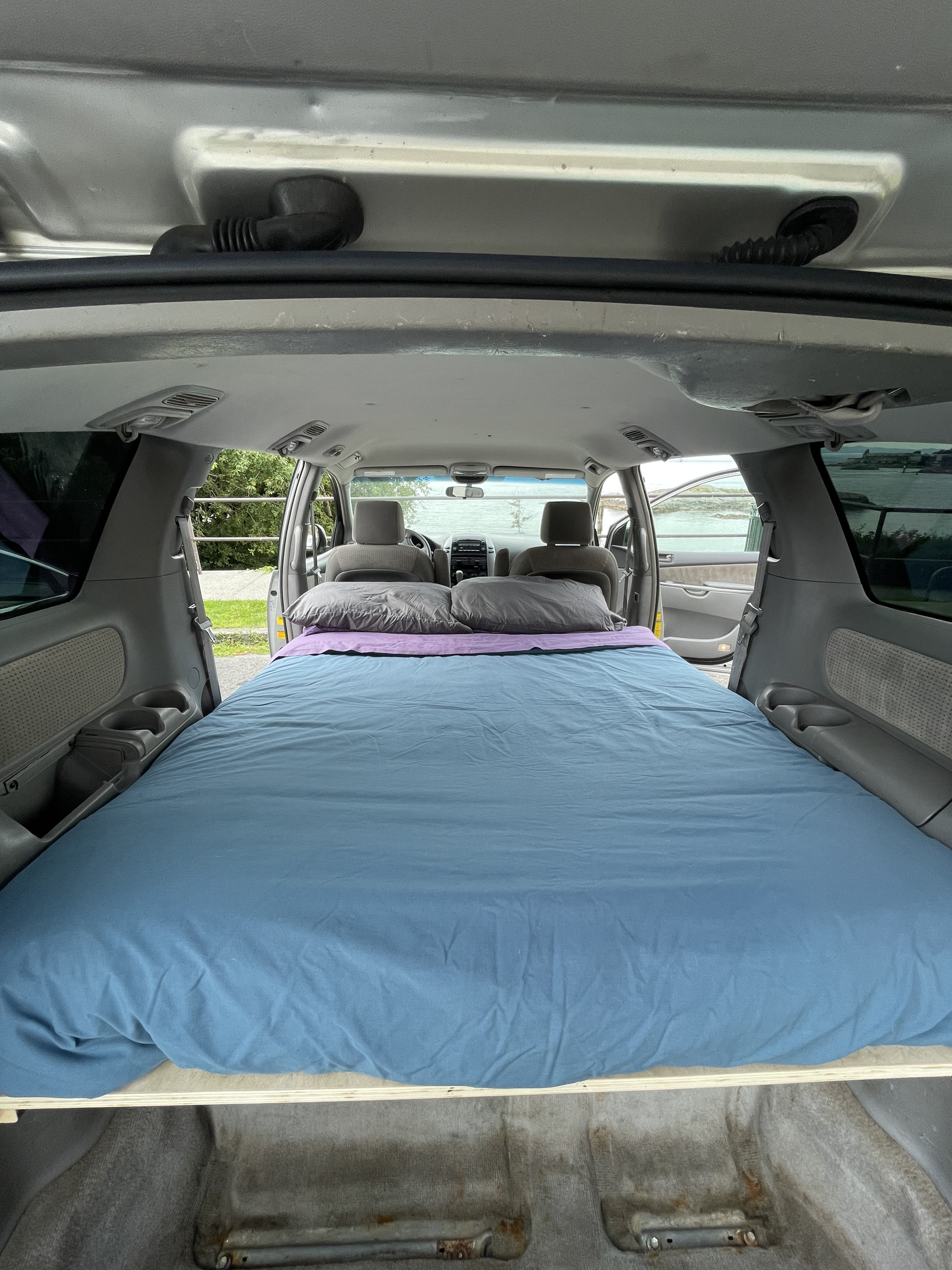 Interior of a converted minivan, showing a blue bed with pillows. Ready for an RV rental adventure or car camping.. Toyota Granvia 2008