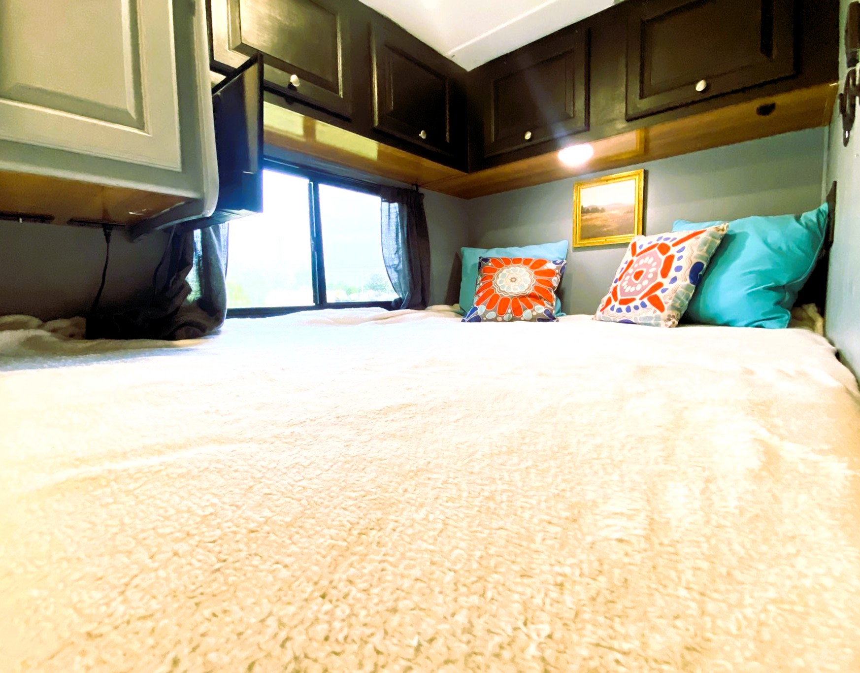 Cozy RV rental bedroom: bed with fuzzy blanket, colorful pillows, dark overhead cabinets, TV, and window.. Coachmen Leprechaun 2016