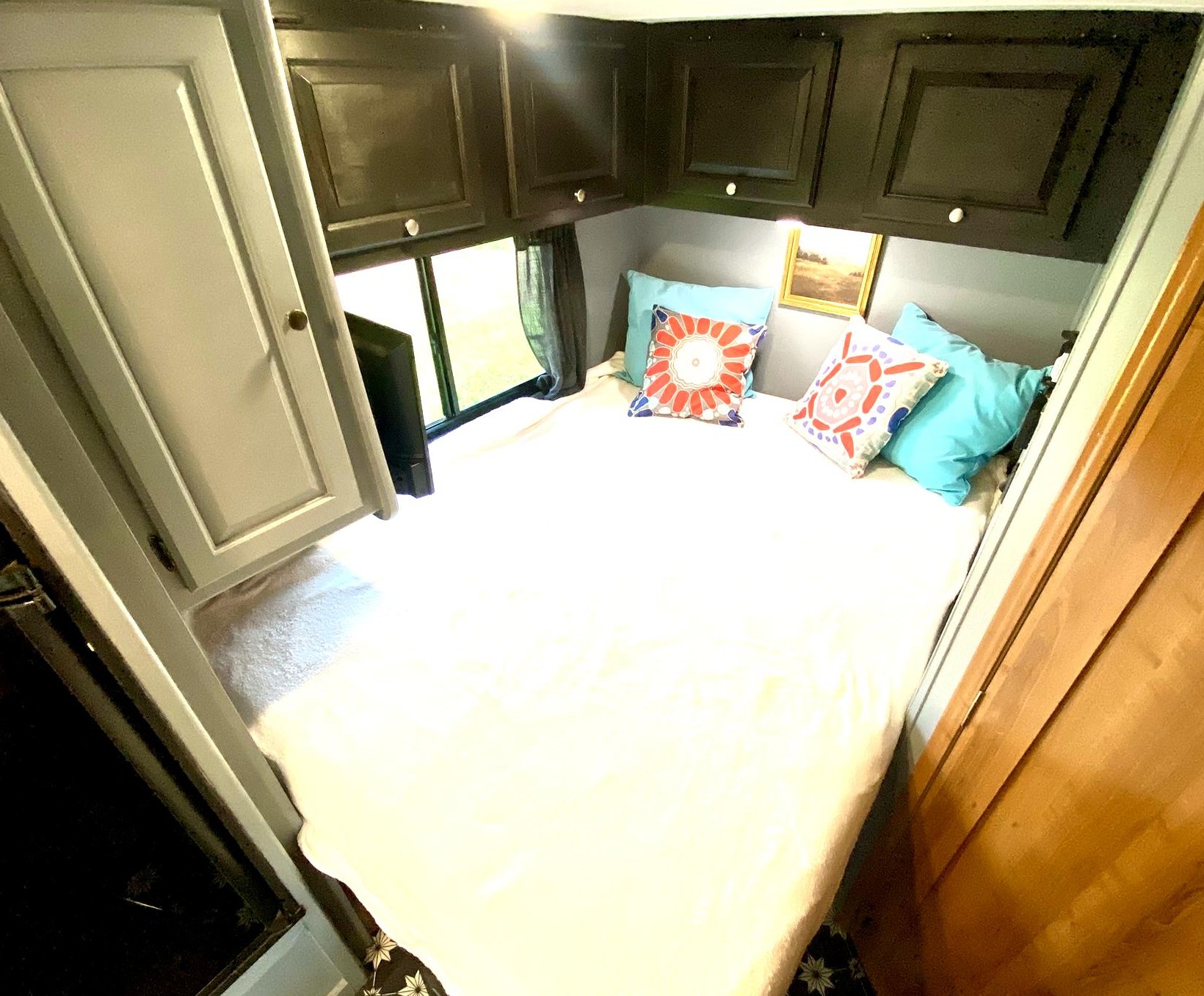 Compact RV rental bedroom featuring a comfortable bed with colorful pillows, dark overhead storage, and a scenic window view.. Coachmen Leprechaun 2016