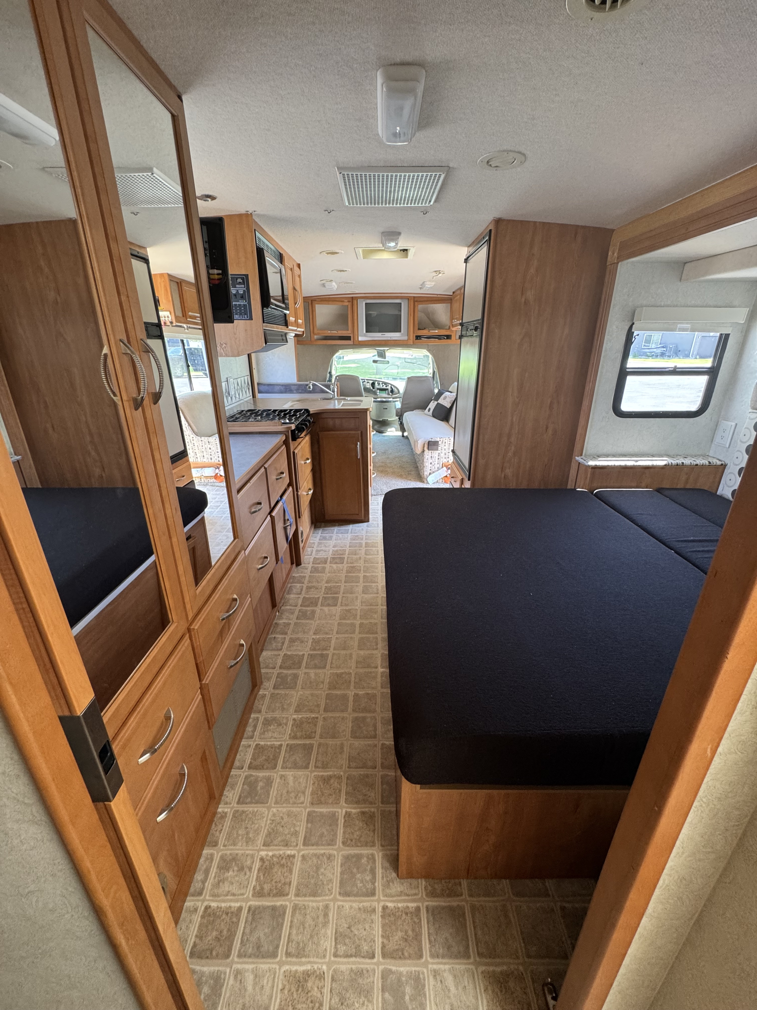 This RV rental interior shows the kitchen, living area with a TV, and a bed. Wood cabinets and patterned floor are visible.. Winnebago Aspect 2006