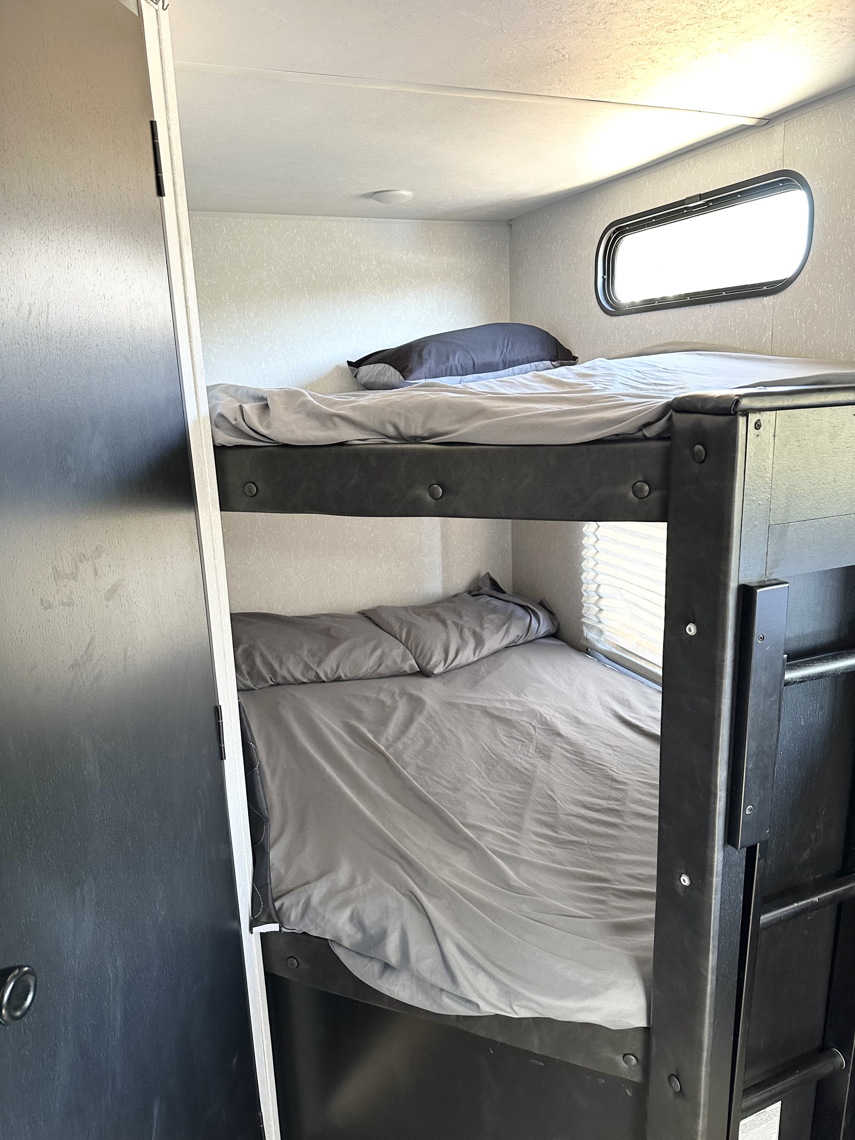 Inside an RV rental: two neatly made bunk beds with grey sheets, a small oval window, and overhead lighting.. Forest River Cherokee Grey Wolf 2021