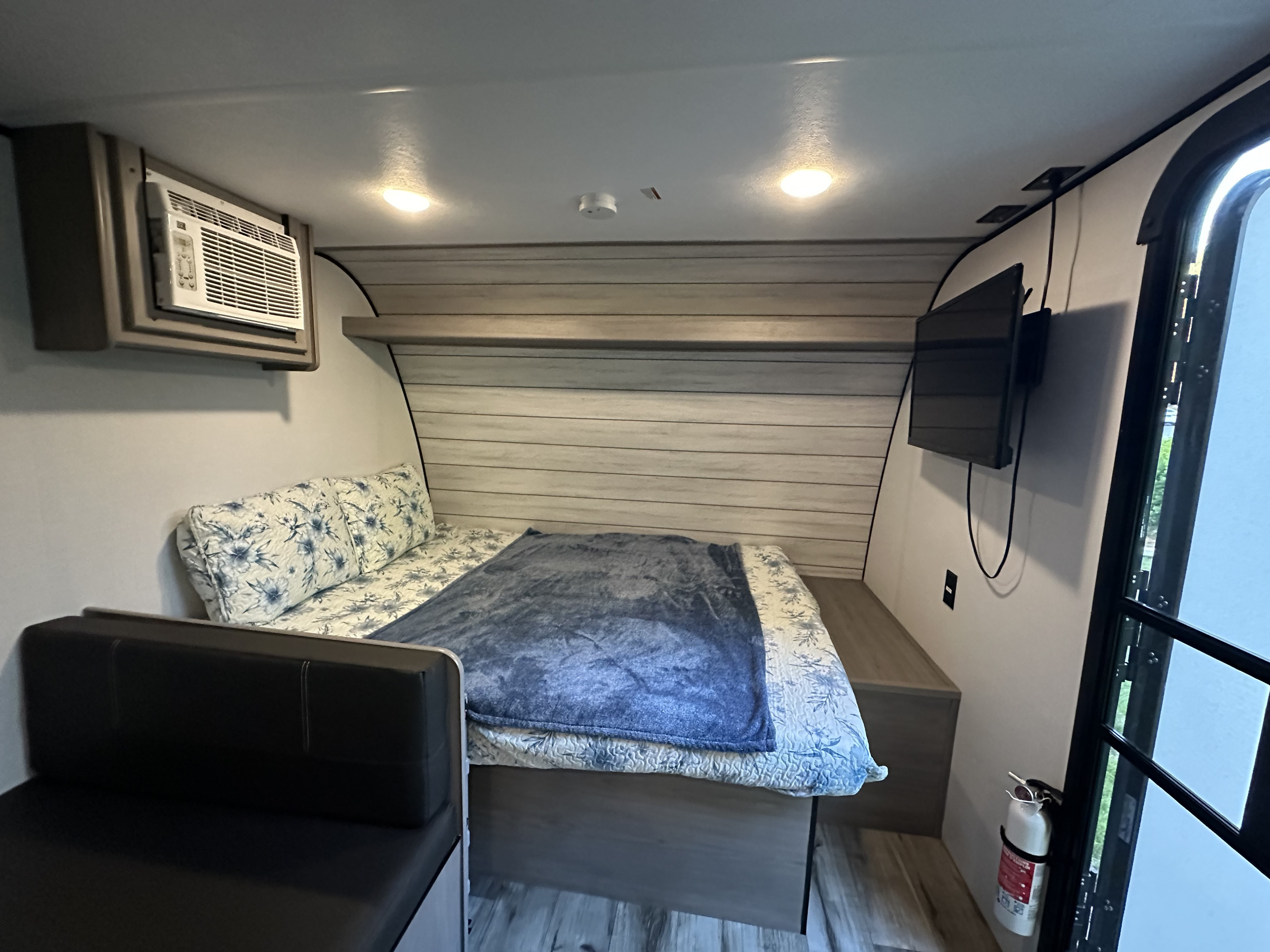 Inside this RV rental: a compact sleeping area with a bed, small couch, AC unit, and wall-mounted TV for your travel comfort.. Dutchmen Coleman lantern 17b 2024