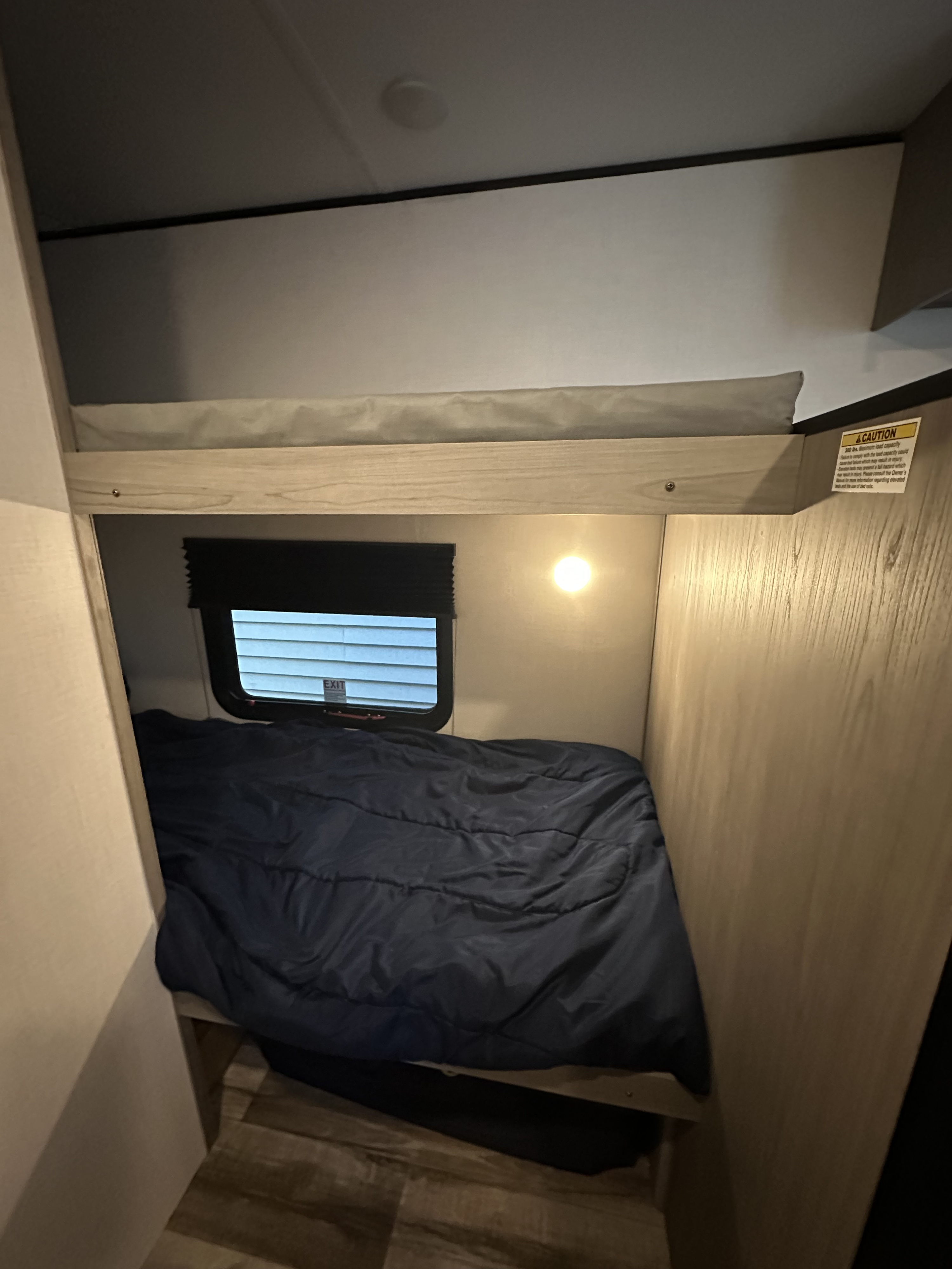 Inside an RV rental, a two-tier bunk bed setup with a window, wall light, and a safety caution on the upper bunk.. Dutchmen Coleman lantern 17b 2024