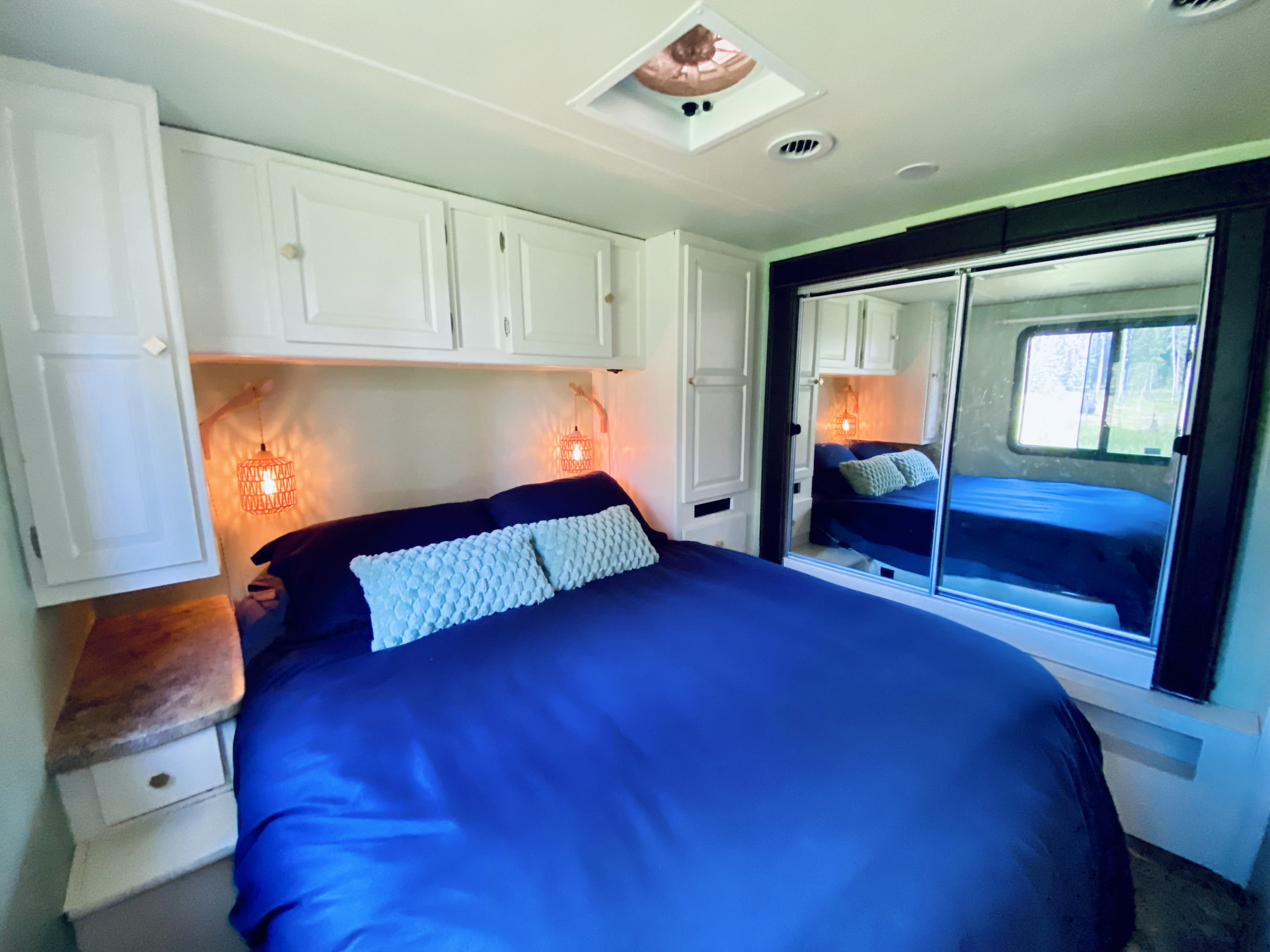Cozy RV rental bedroom features a blue bed, white overhead cabinets, warm accent lights, and mirrored closet.. Coachmen Leprechaun 319 DS 2015