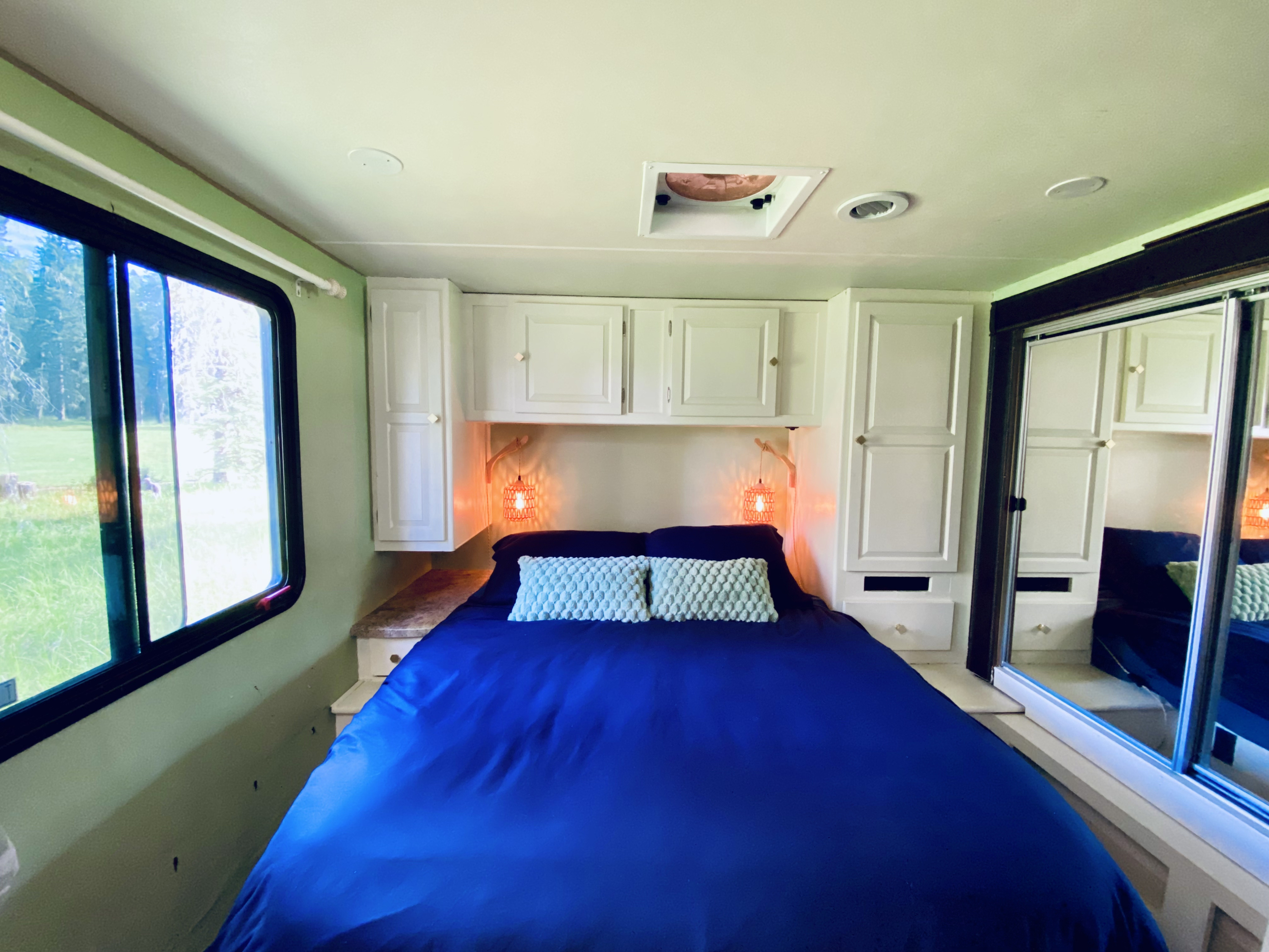 Cozy RV rental bedroom featuring a blue bed, built-in white storage, bright window views, and warm decorative lighting.. Coachmen Leprechaun 319 DS 2015