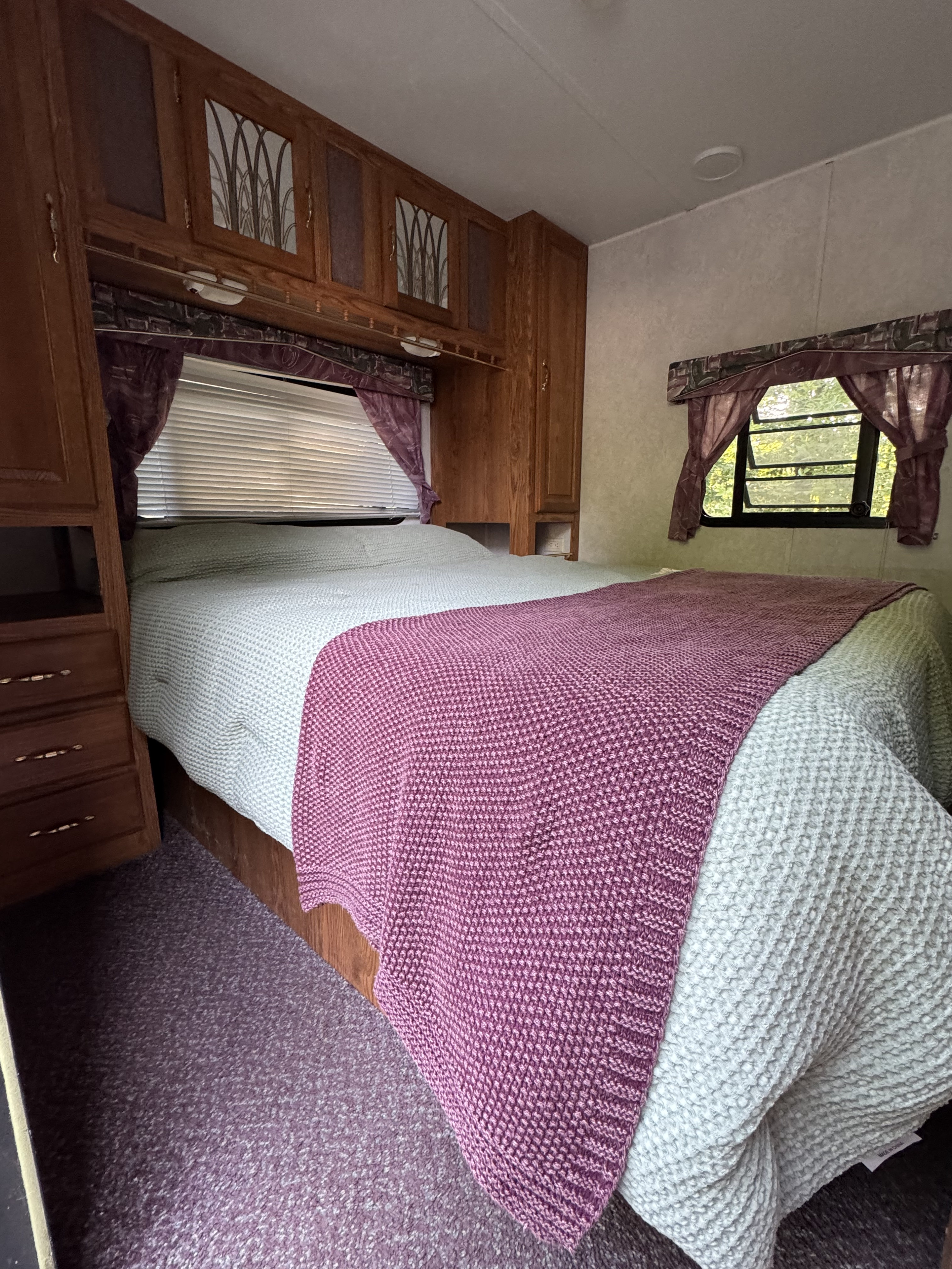 RV rental bedroom: a cozy bed with purple and grey blankets, wooden cabinetry, and a window with tree views.. Forest River Wildwood 2000