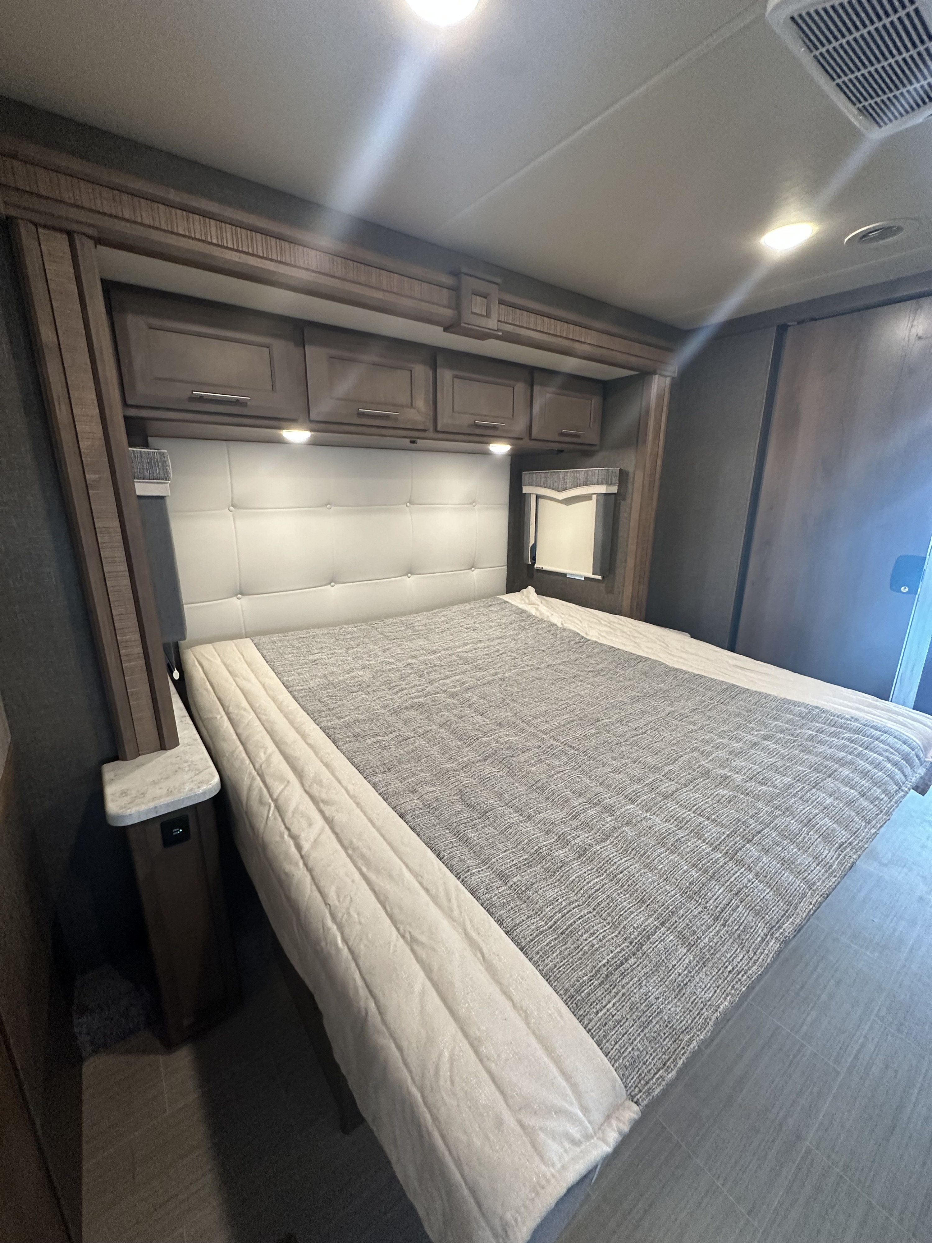 This RV rental's bedroom features a comfy bed with a grey textured blanket, padded headboard, and sleek overhead storage.. Thor Challenger 2022