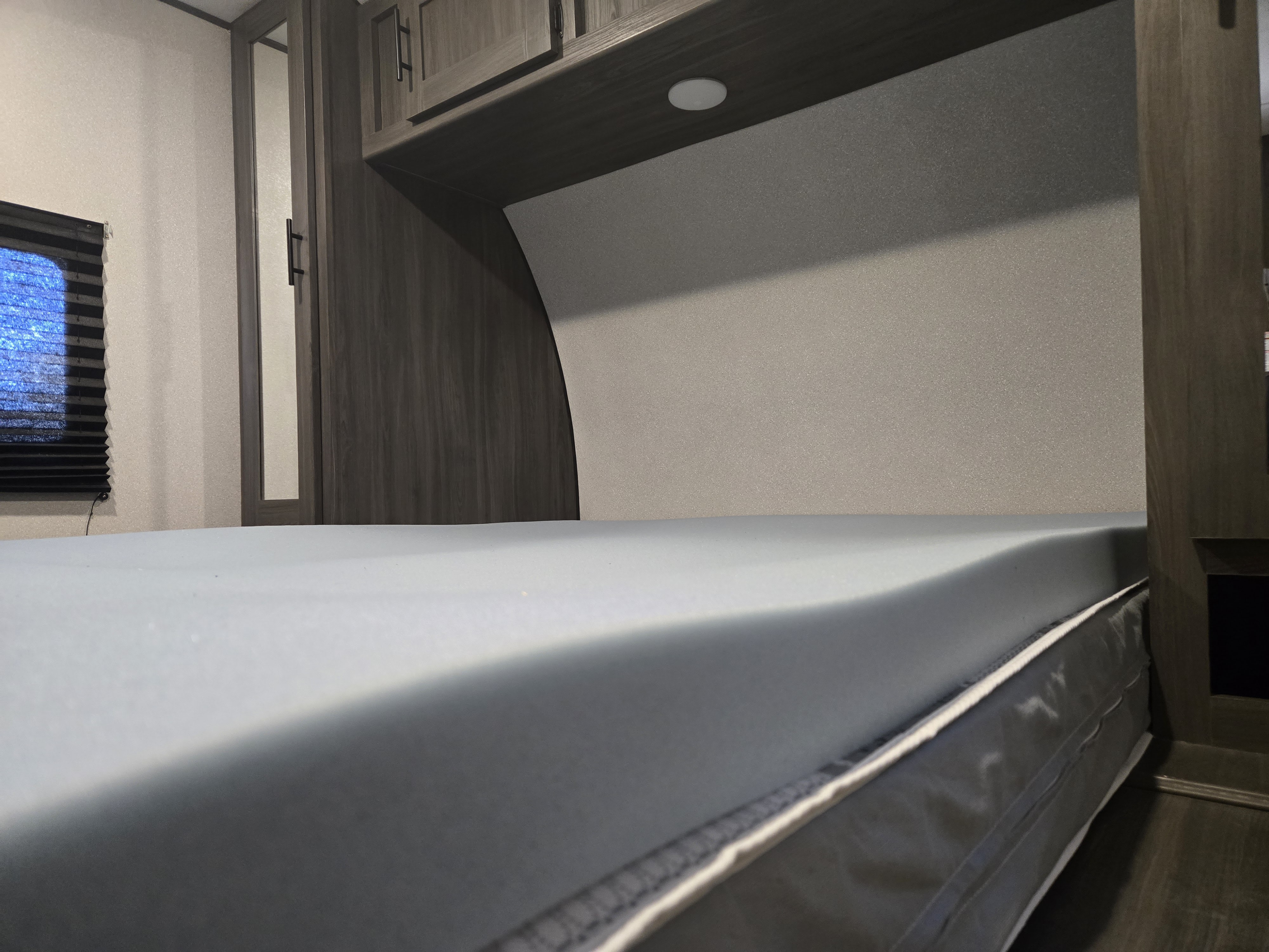 View of an RV rental bed with a light grey foam mattress topper, ample overhead storage, and a window.. Forest River Shasta 2021