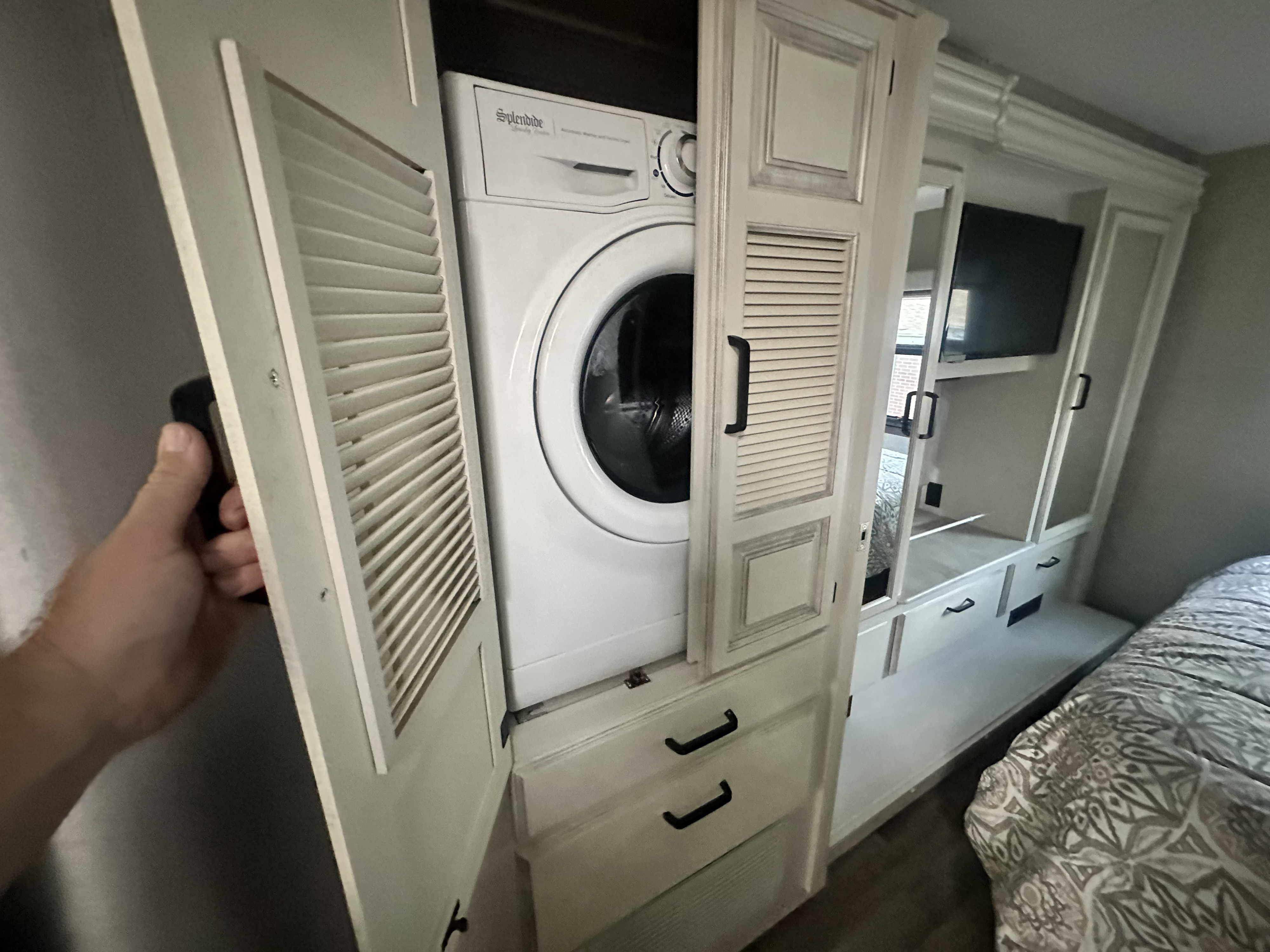 Inside an RV rental, a hand opens a louvered door revealing a compact Splendide washer, next to a bed and storage.. Jayco Precept Prestige 2022