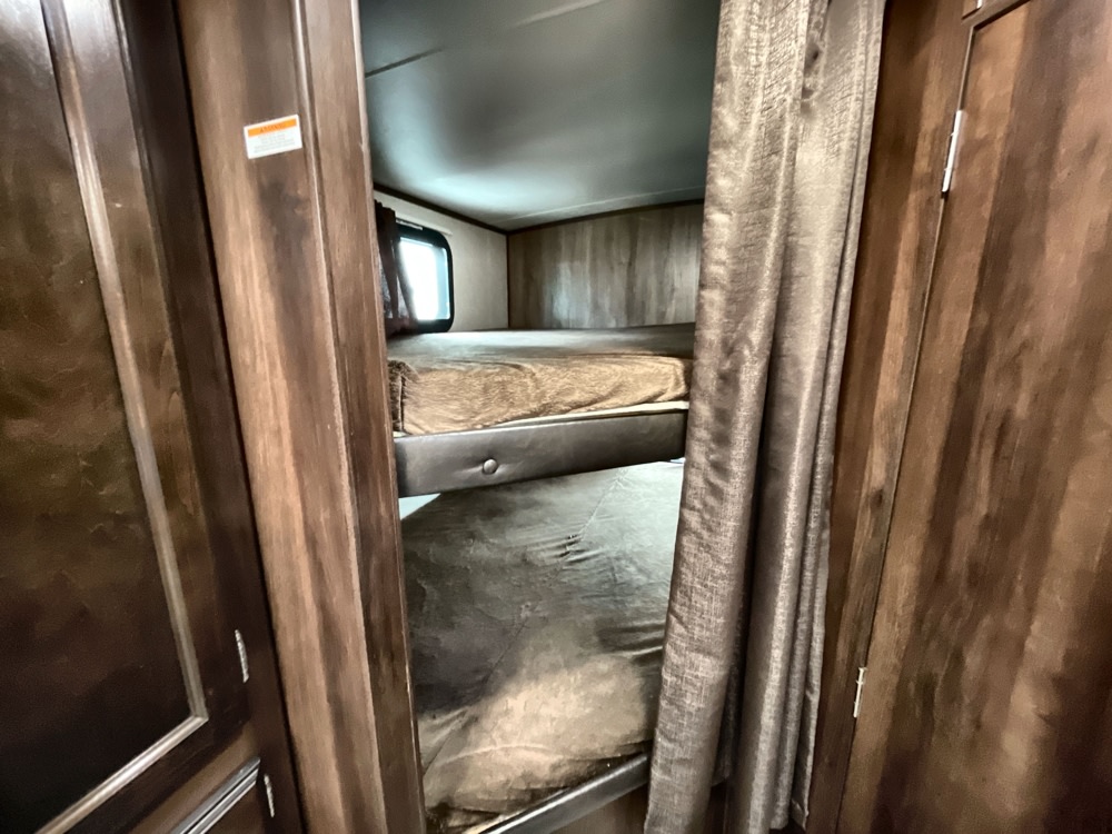 Inside an RV rental: two comfy bunk beds with mattresses and a window. Features cozy wood paneling and a privacy curtain.. Jayco White Hawk 2019