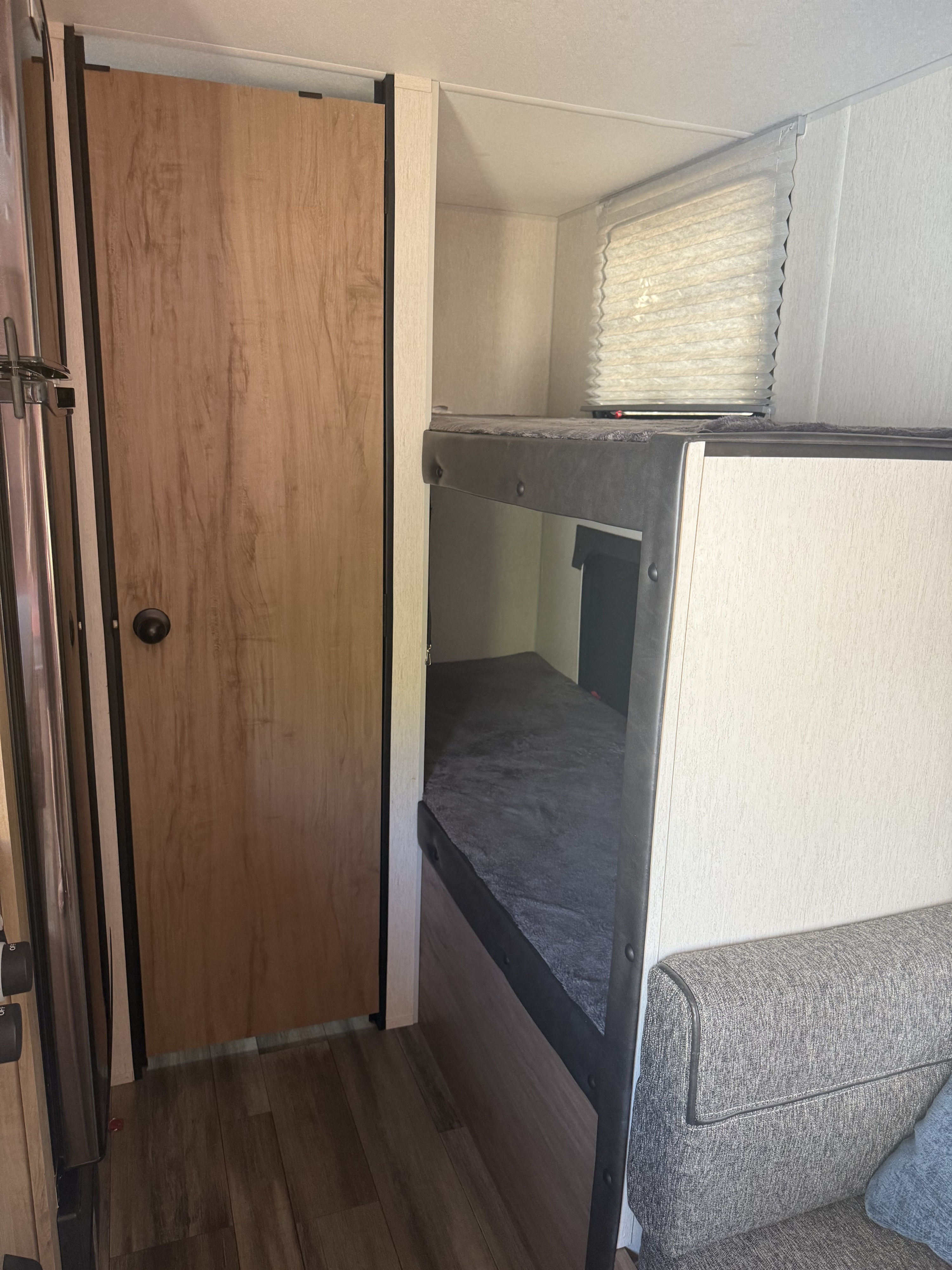 Cozy RV rental interior with a wooden door, two bunk beds, a window with blinds, and a comfortable grey sofa.. Forest River Cherokee Wolf Pup 2023