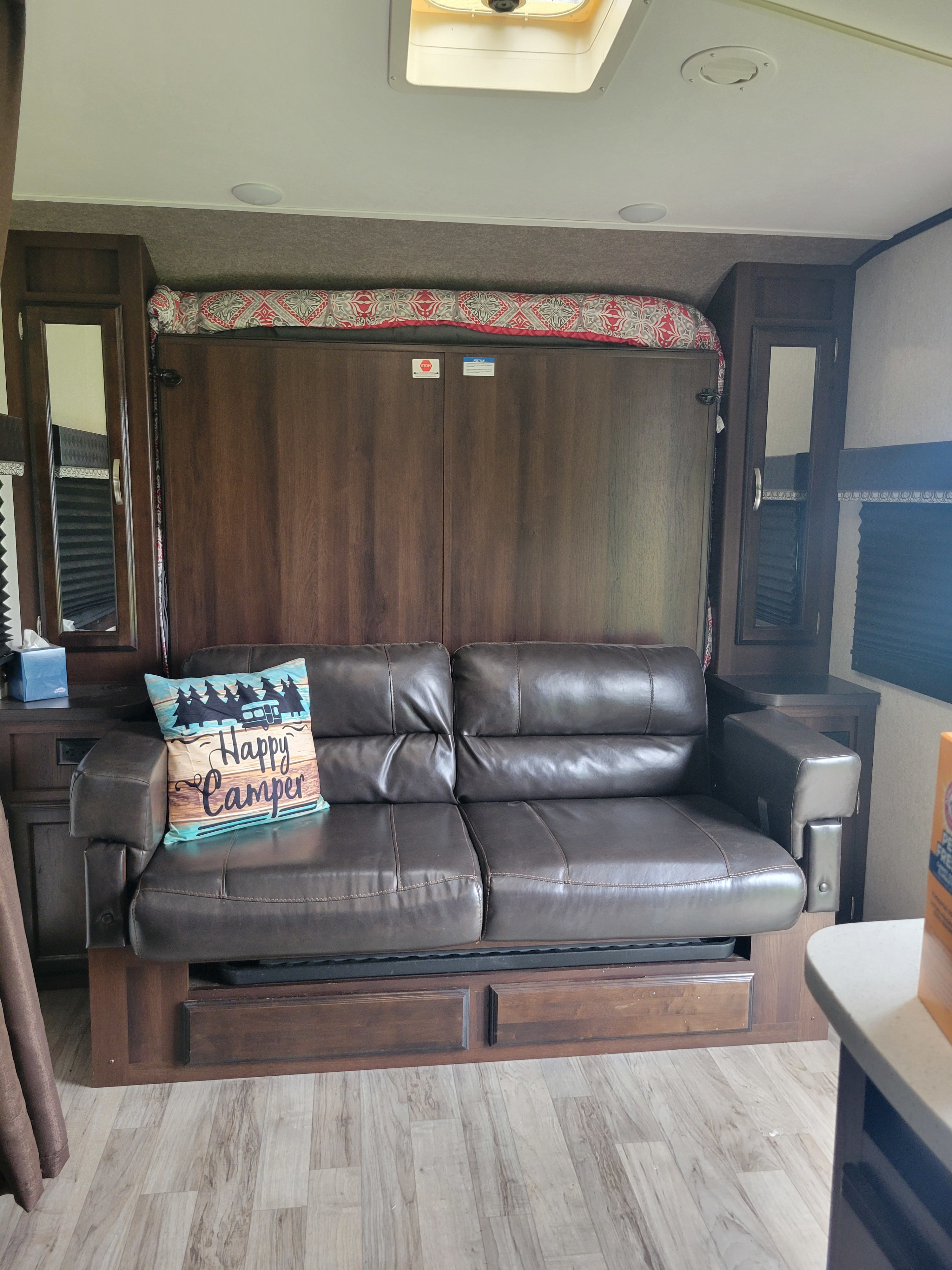 Inside an RV rental: a brown leather sofa with a "Happy Camper" pillow and a cleverly hidden murphy bed above.. Jayco White Hawk 2019