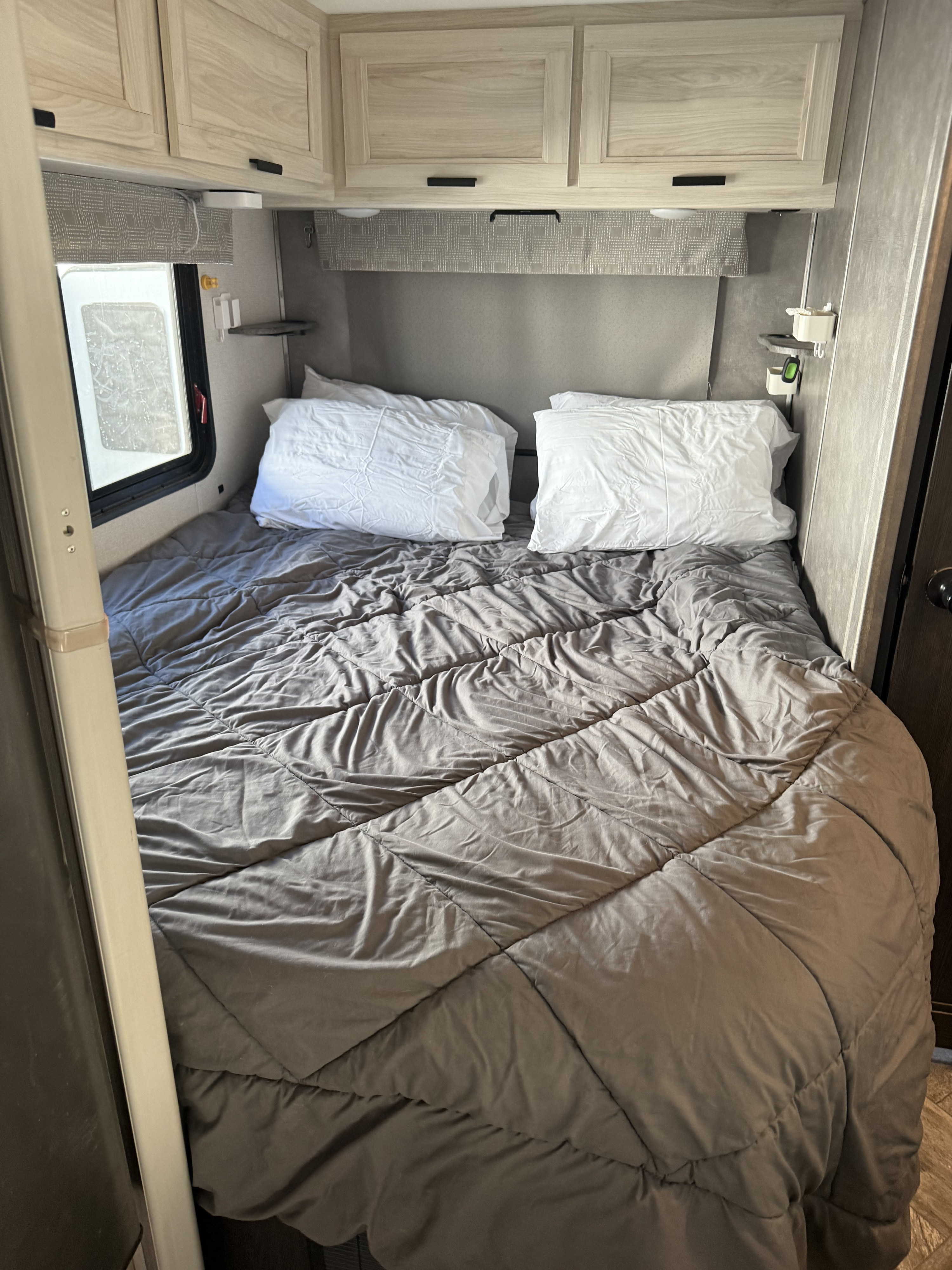 RV rental bedroom: a made bed with grey comforter & white pillows, overhead wood cabinets, and a window for light.. Forest River Sunseeker LE 2022