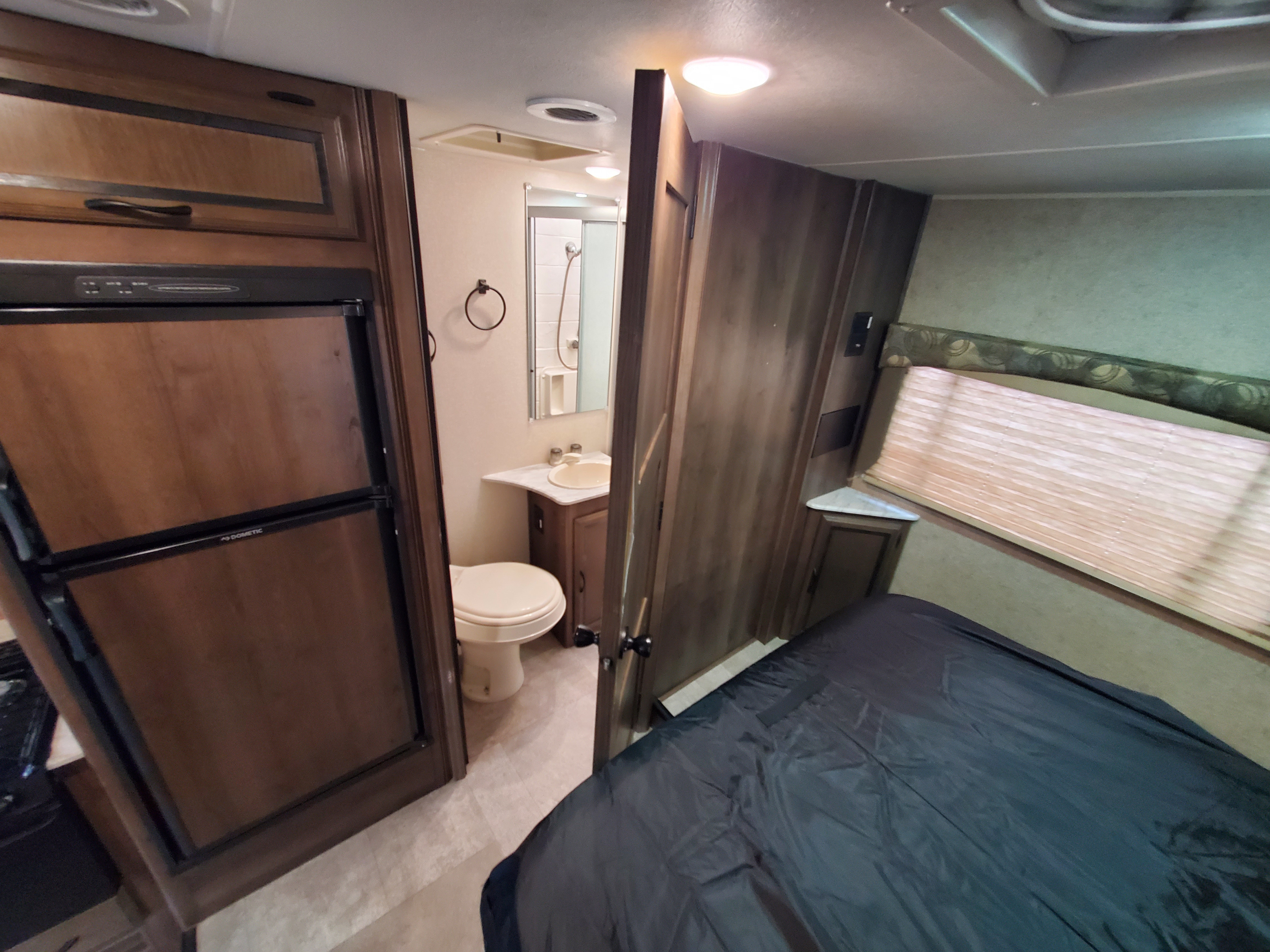 RV rental interior featuring a compact bedroom with a dark bed, an adjacent bathroom with toilet, sink, shower, and a refrigerator.. Coachmen Freelander 2018