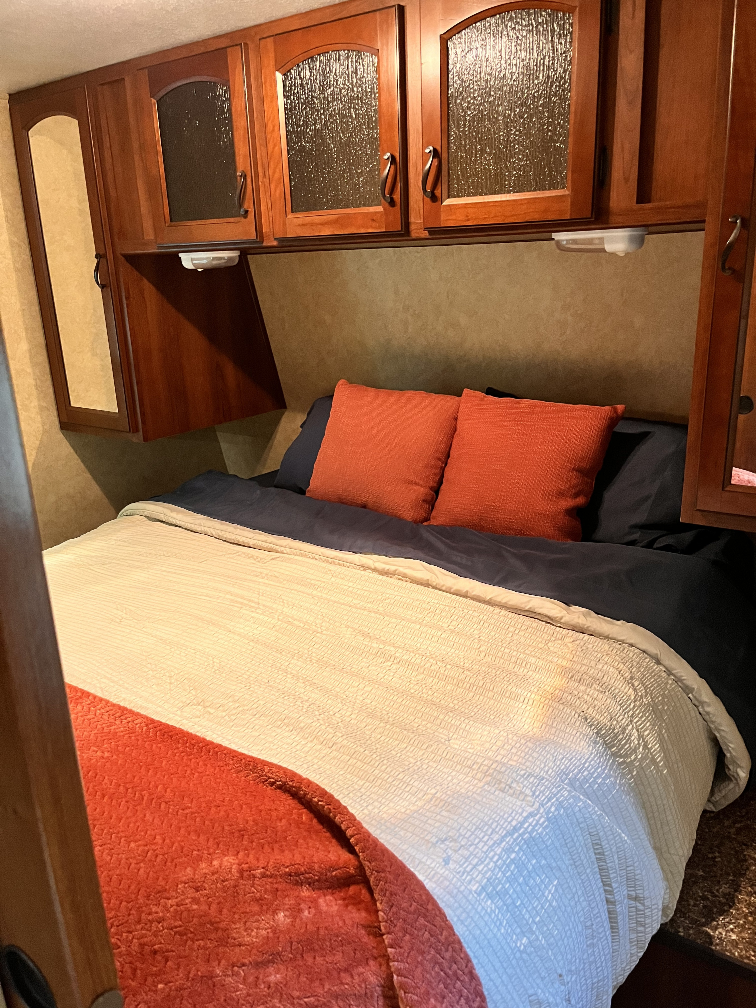 Compact RV rental bedroom with a bed, blue sheets, orange pillows, and wooden overhead cabinets.. Forest River Lacrosse Luxury Lite 2012