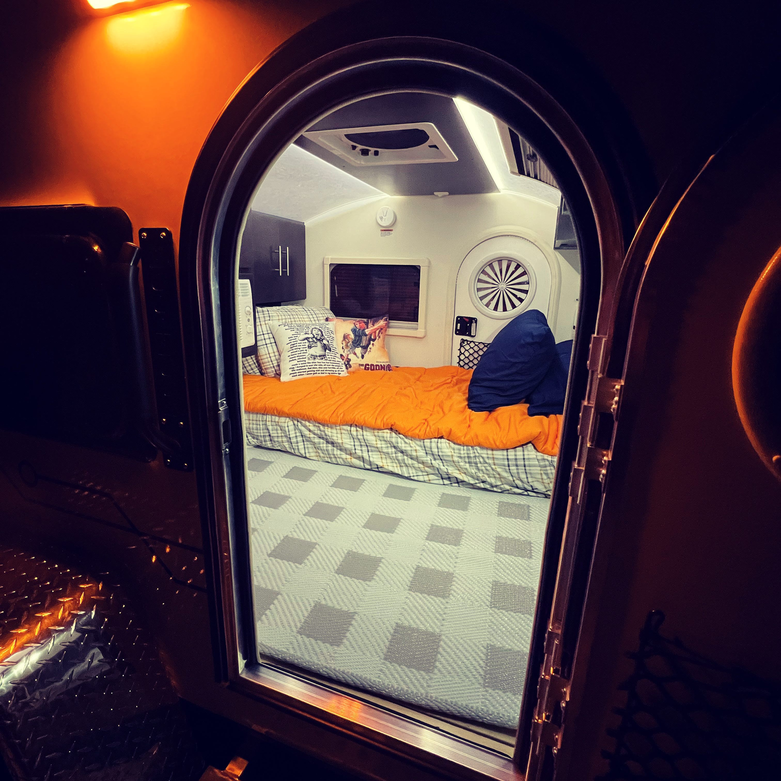 Inside this compact RV rental: a cozy bed with Goonies pillow, window, and warm lighting for adventures.. nuCamp T@G 2021